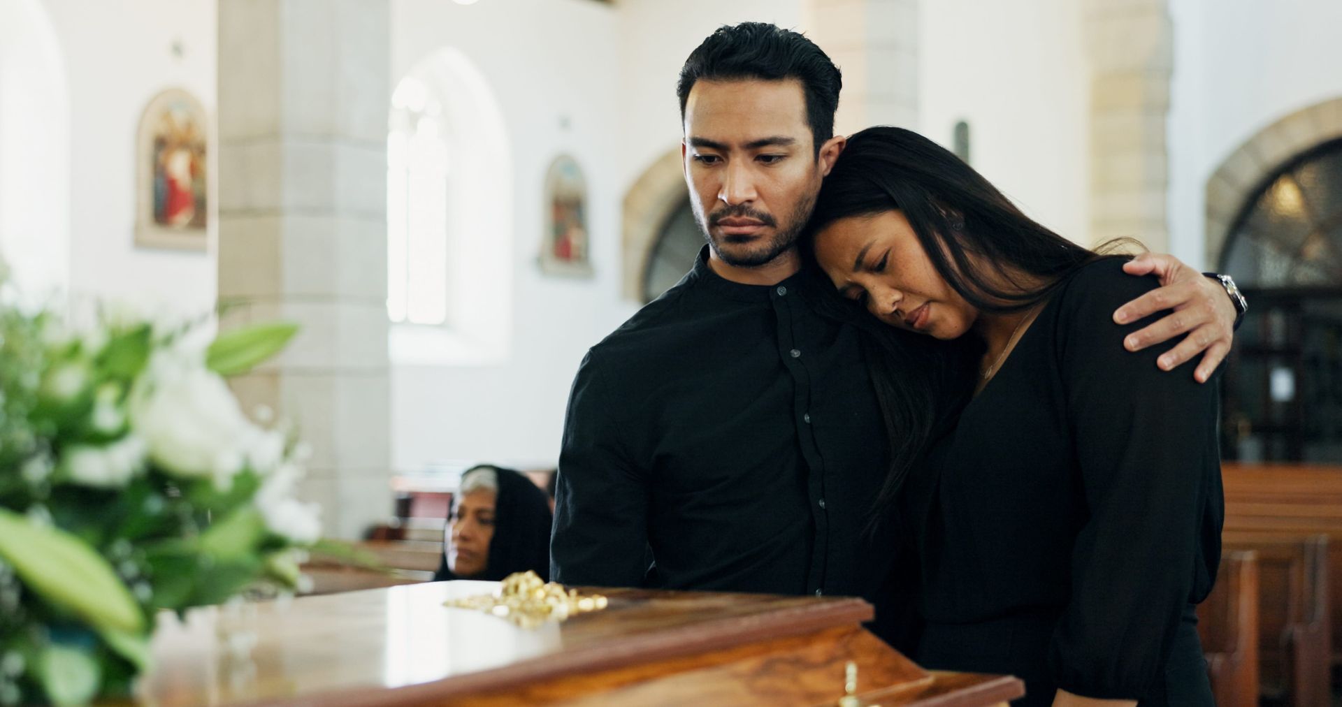 Two people in black clothing stand in a church, leaning against a wooden casket with somber expressions.