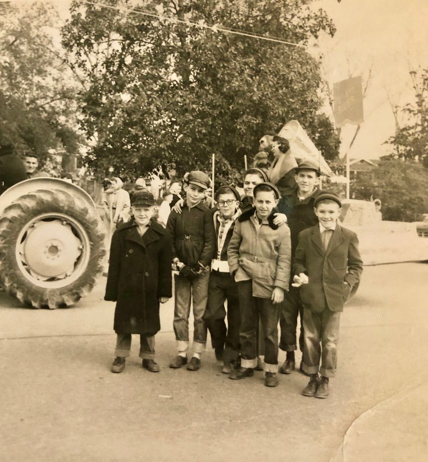 Group of boys posing near a tractor and a tree. They wear jackets, hats, and stand on pavement in an outdoor setting.