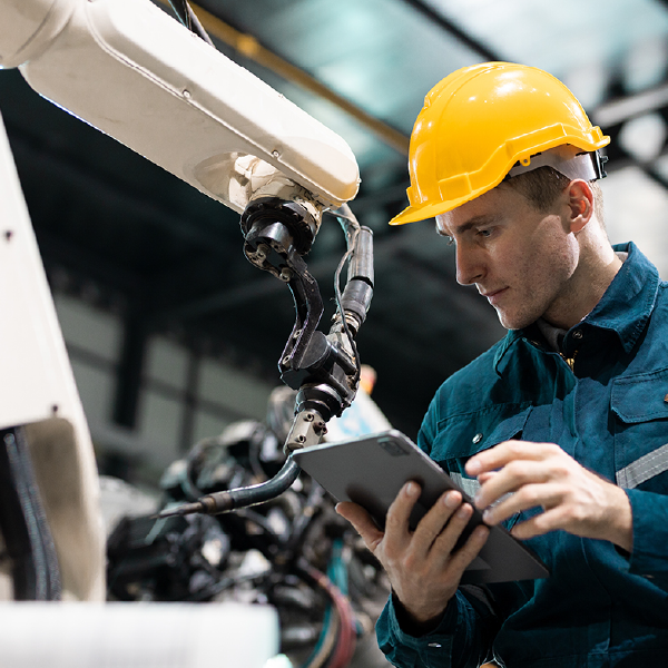 Technician in yellow hard hat checks industrial robot arm with a tablet in a factory