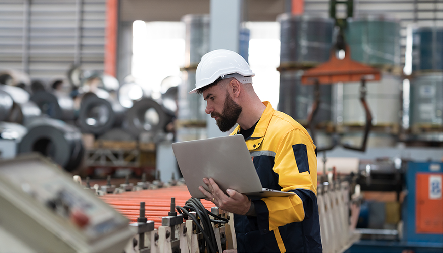 Engineer in hard hat using laptop at a factory workstation, inspecting equipment.