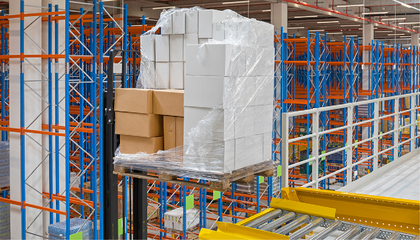 A forklift in a warehouse raises a pallet stacked with white and brown cardboard boxes wrapped in clear plastic.