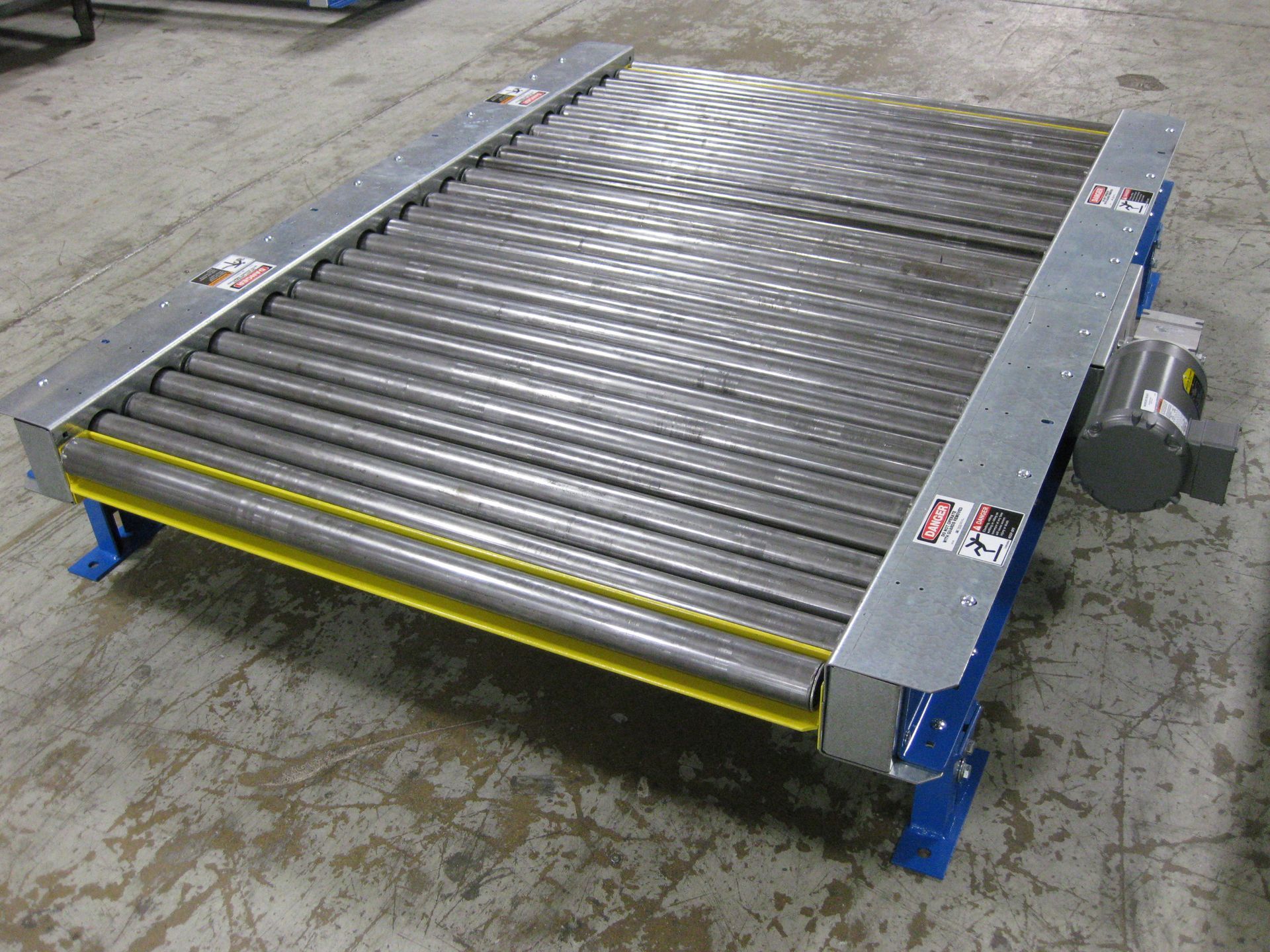 Industrial roller conveyor on a concrete floor, with metal rollers, blue supports, and yellow safety edges