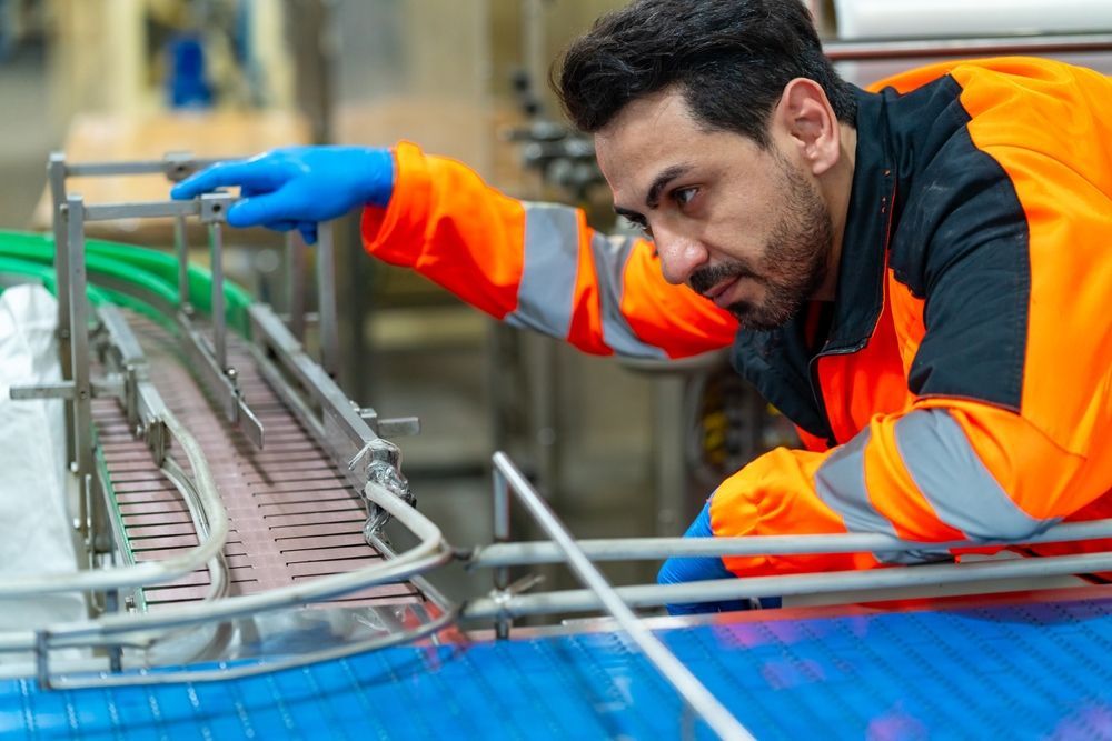 Worker in orange safety jacket and blue gloves inspecting industrial machinery on a factory line
