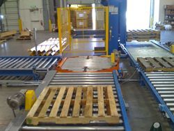 Industrial pallet wrapping machine with conveyor belts and safety fencing.