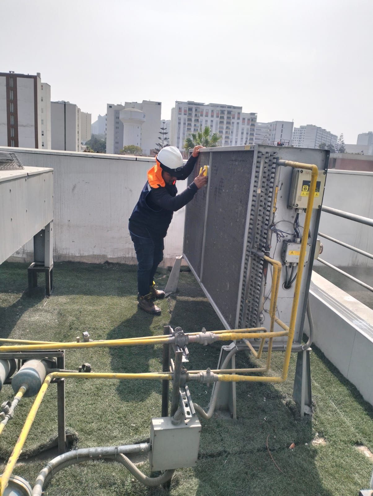 Un trabajador con casco inspecciona la unidad de climatización de la azotea, junto a una barandilla de seguridad amarilla.