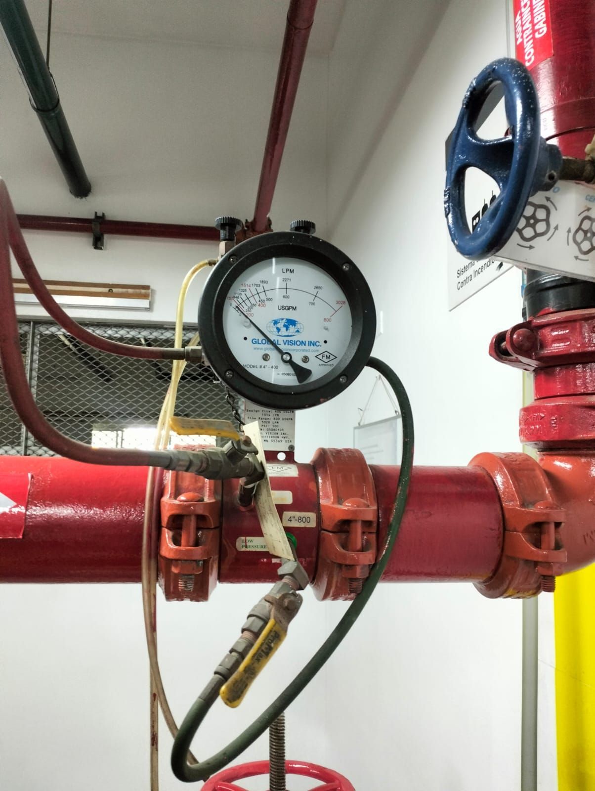 Manómetro y tubería roja de extinción de incendios con rueda de válvula azul en una sala industrial.