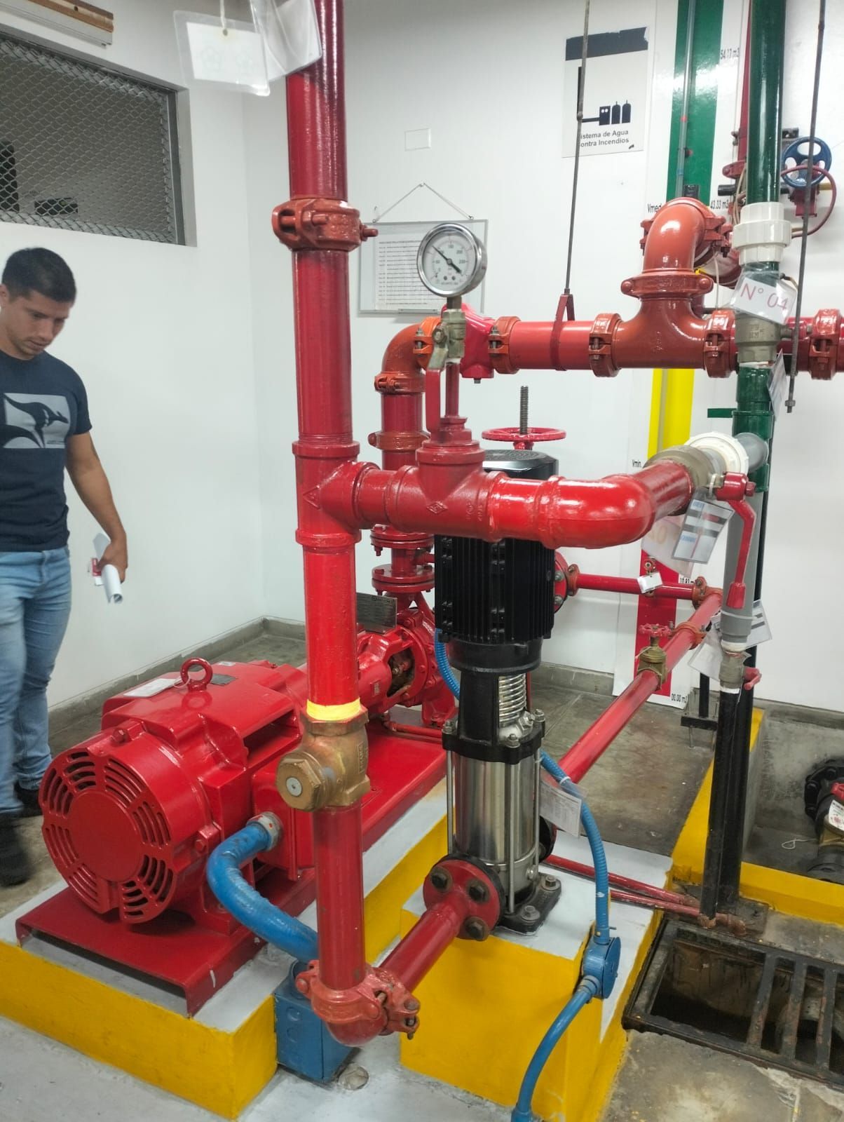 Bomba industrial roja con tuberías y manómetros rojos en un cuarto de servicio; un hombre está de pie junto a ella.
