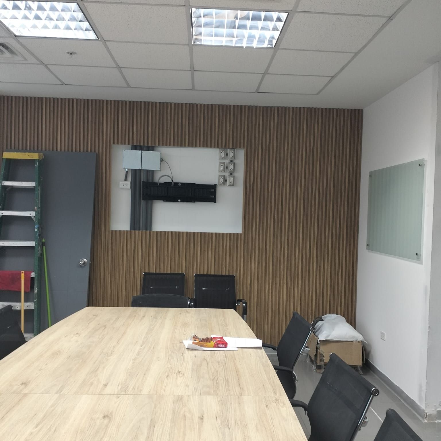 Pequeña sala de reuniones de oficina con una mesa grande, sillas, televisor montado en la pared, estantes y pizarra blanca.