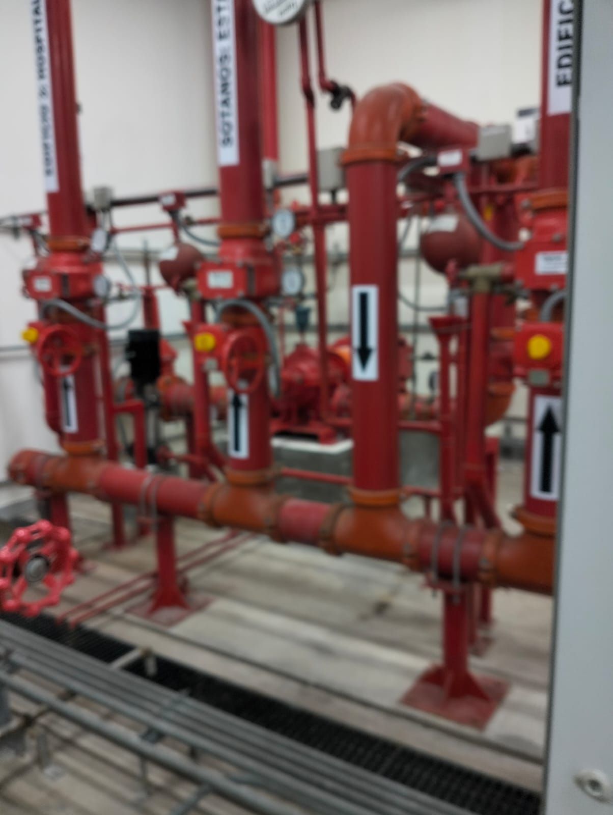 Tuberías y válvulas rojas de rociadores contra incendios en una sala de máquinas, con flechas direccionales blancas.