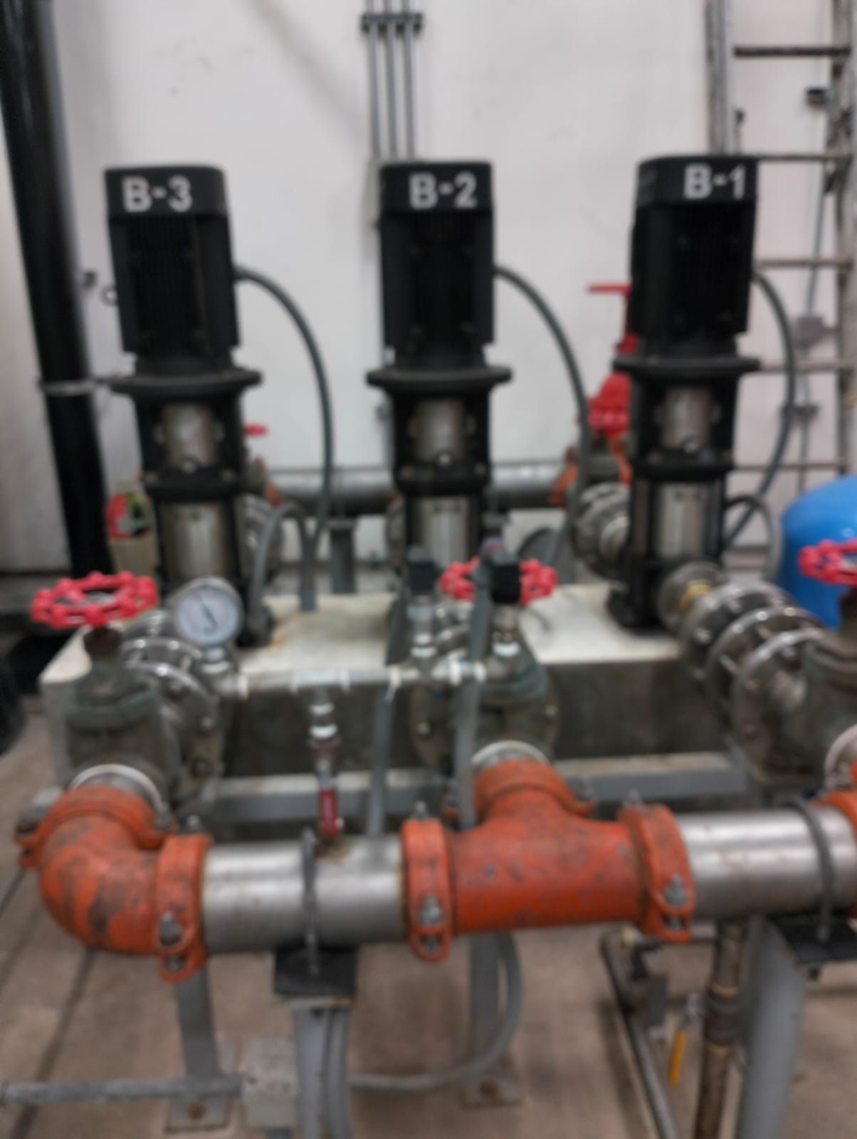 Tubería industrial con tres válvulas negras etiquetadas y secciones de tubería naranjas en una sala de máquinas.