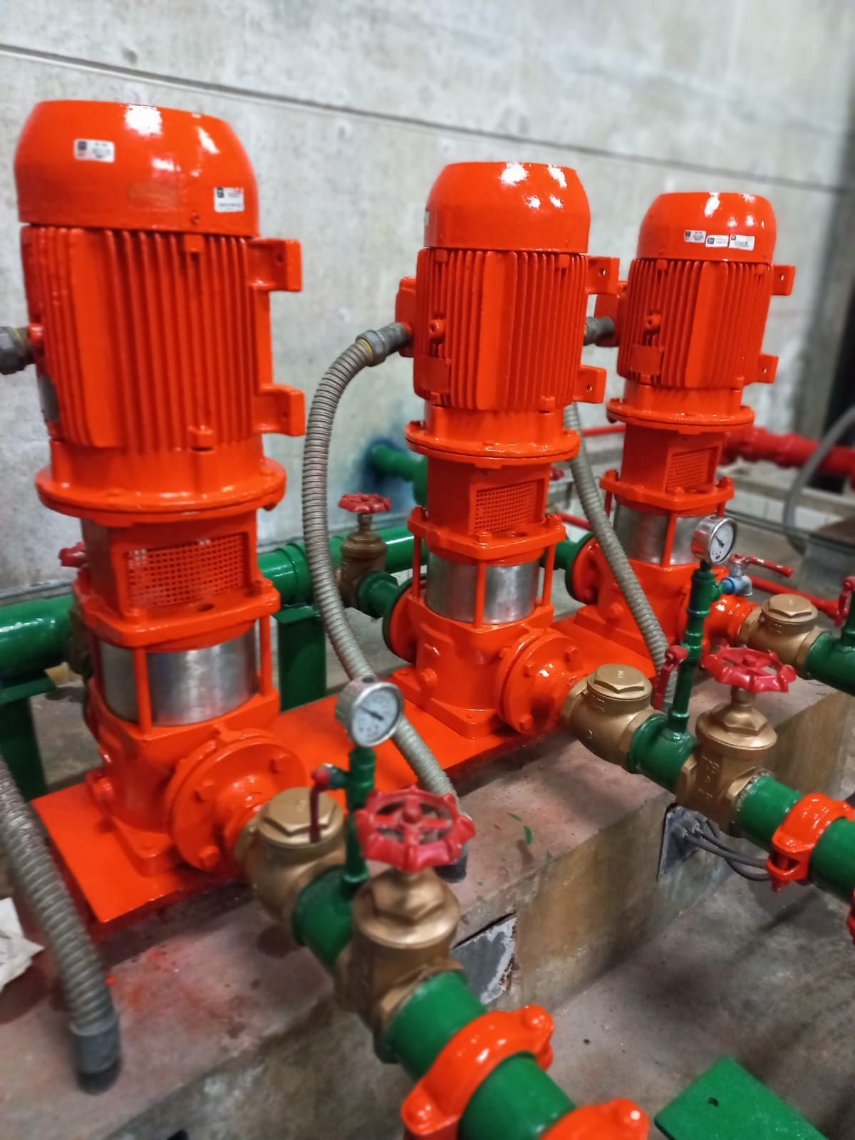Tres bombas industriales de color naranja brillante con tuberías verdes en una sala de máquinas.