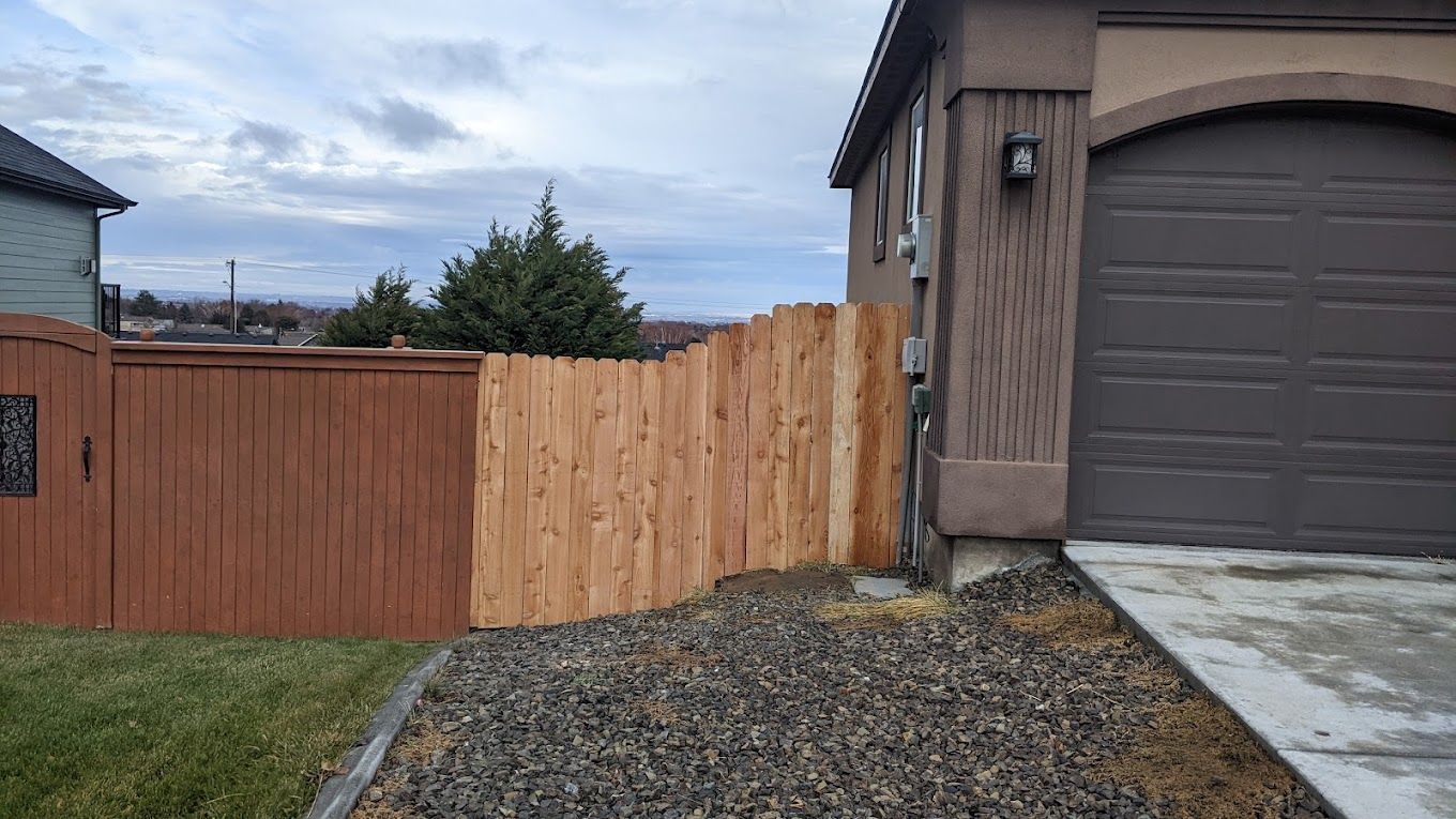 Brown fence next to a garage, on a gravel bed and lawn, under a cloudy sky.