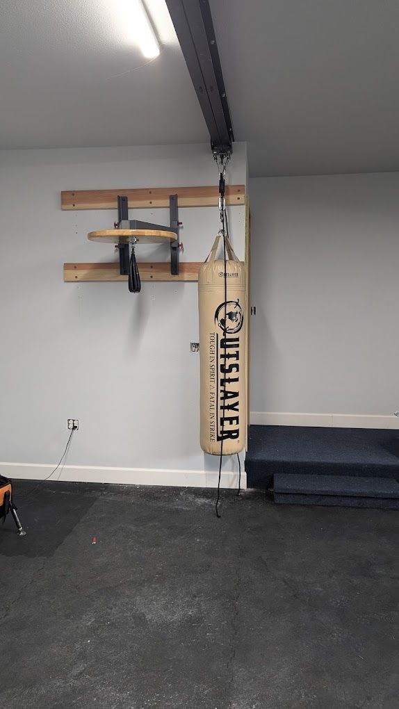 Boxing setup in a gym. A hanging heavy bag, speed bag platform, and pull-up bar on a gray wall. Black floor.