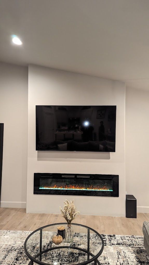 Modern living room: TV above fireplace, black framed. Coffee table with flowers. Black subwoofer.