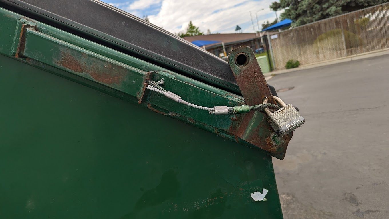 Green dumpster with a rusty metal lid secured with a lock.