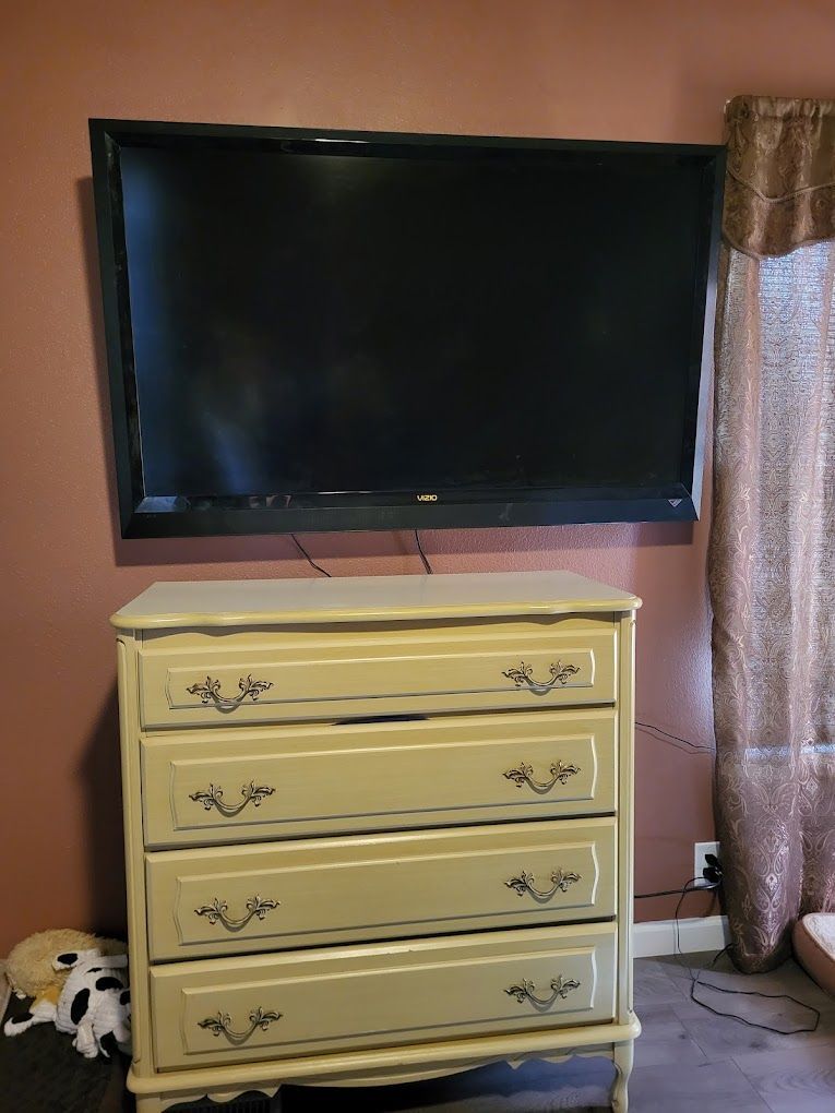 A TV mounted above a beige dresser against a maroon wall. A window with a curtain is on the right.