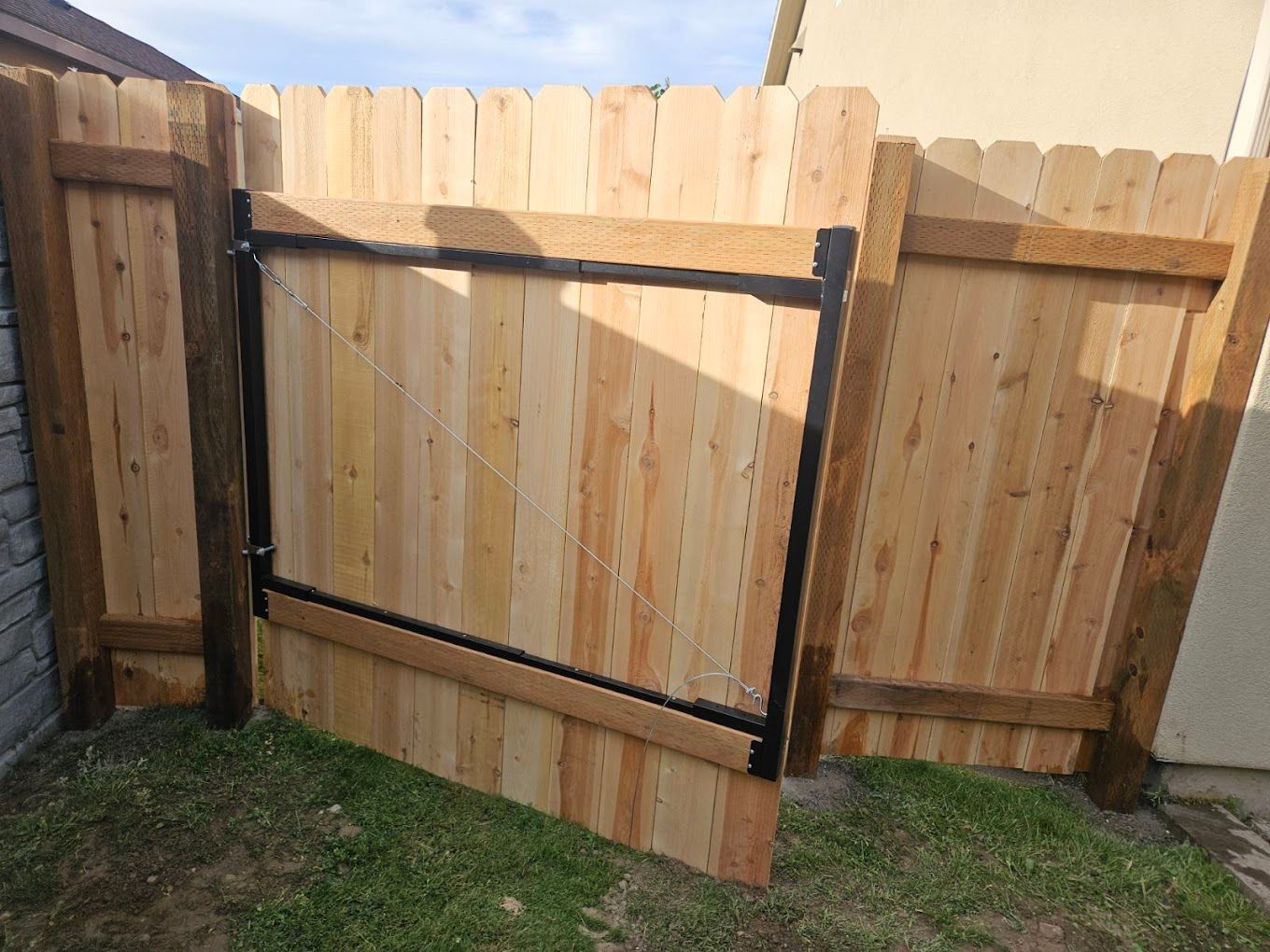Wooden gate ajar in a wooden fence with black metal framing. Gate is on green grass.
