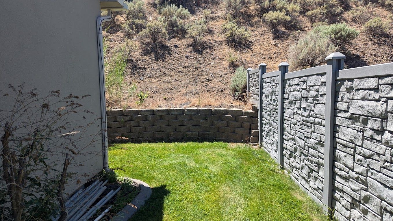 Small backyard with green grass, stone and concrete fences, and a hill in the background.