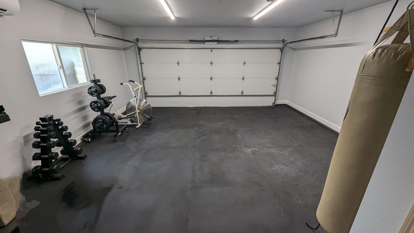 Garage gym with weights, punching bag, and exercise equipment. Dark floor, white walls and garage door.