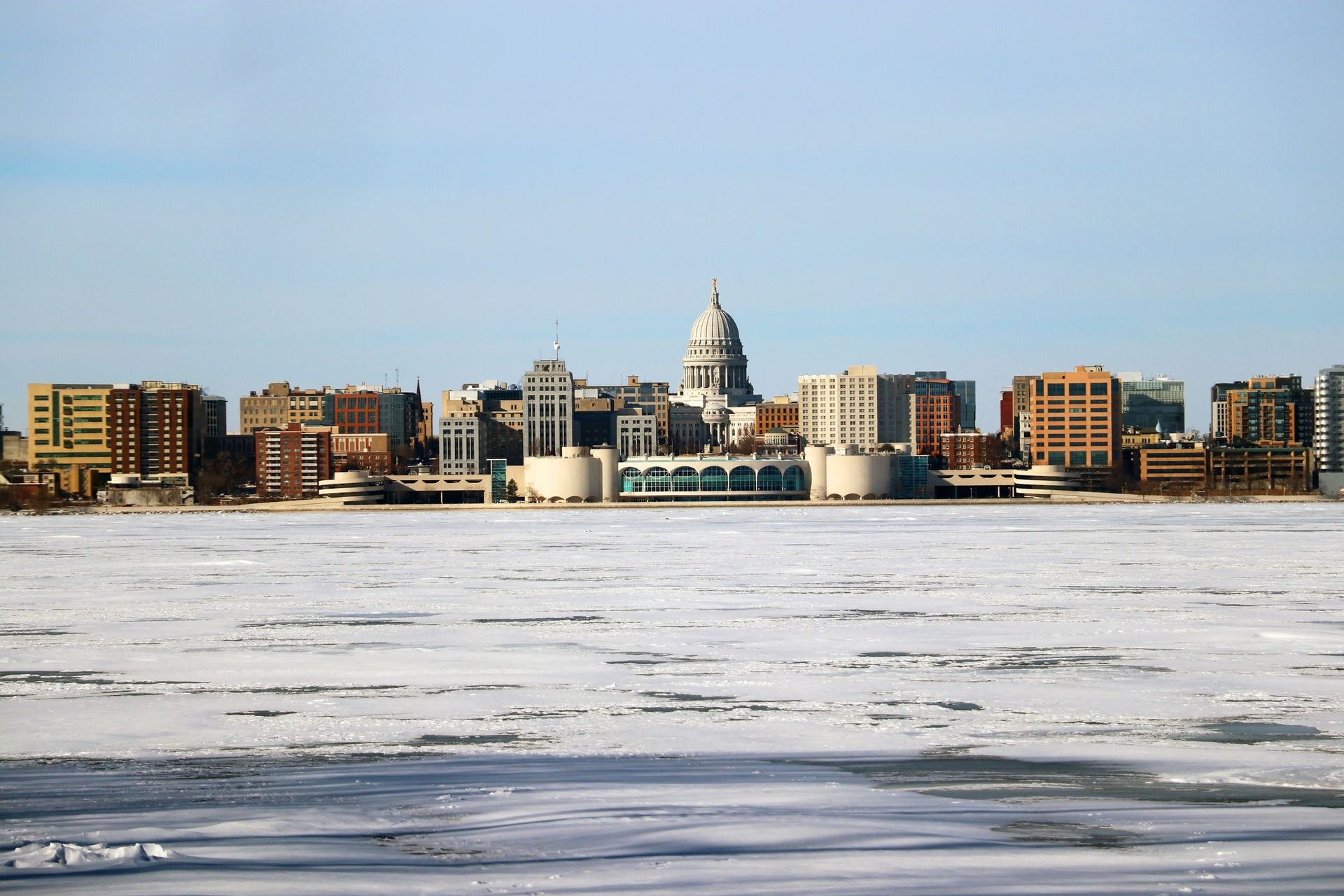 Winter skyline of downtown Madison, Wisconsin, featuring the Wisconsin State Capitol and Monona Terrace Convention Center viewed across frozen Lake Monona.