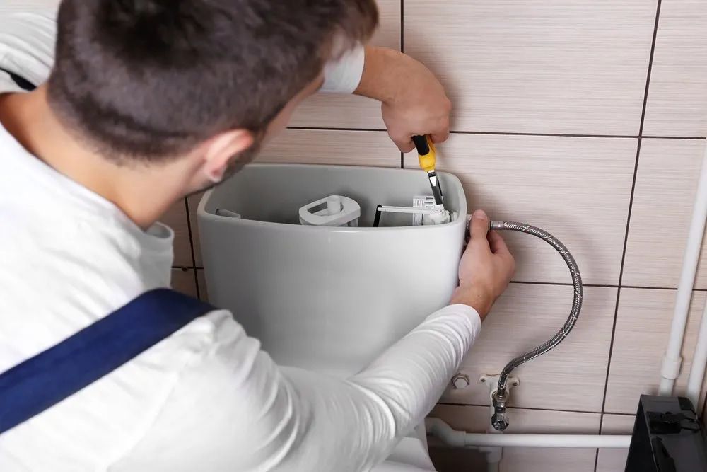 A man is fixing a toilet in a bathroom with a screwdriver — Malouf Plumbing In Pie Creek, QLD