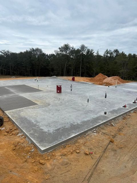 Concrete foundation slab at a construction site with pipes, dirt piles, and trees in the background
