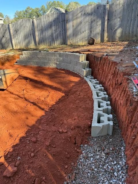 Curved cinder block retaining wall under construction along a red dirt slope beside a wooden fence