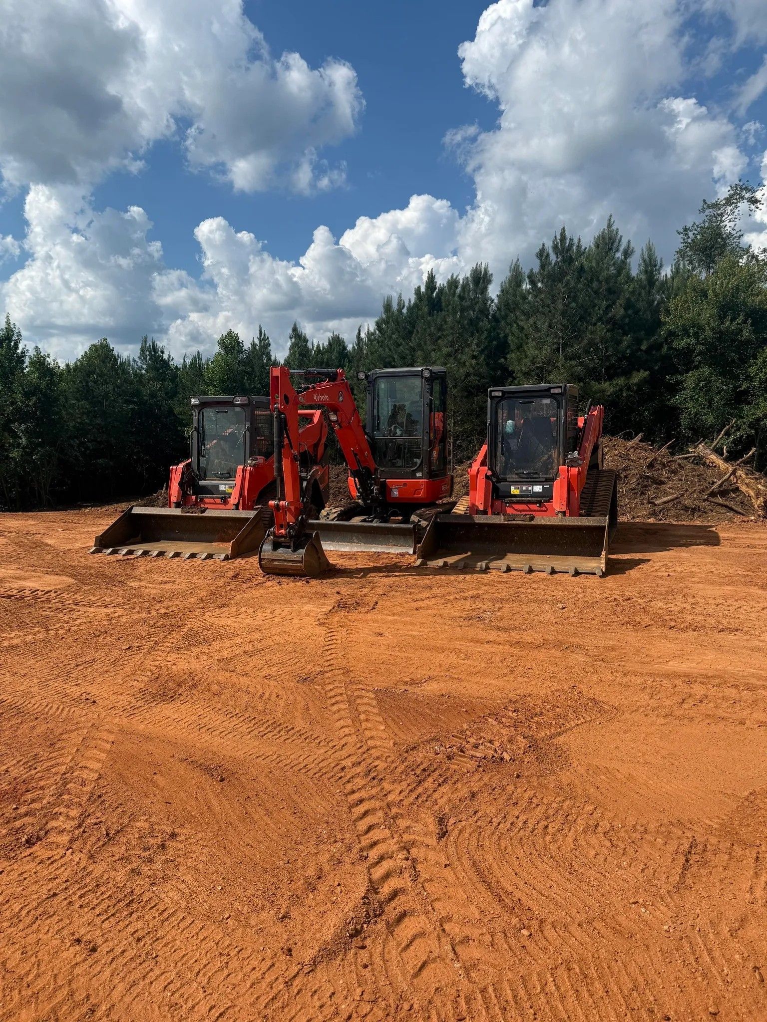 Three red excavators parked on a muddy dirt lot under a cloudy sky