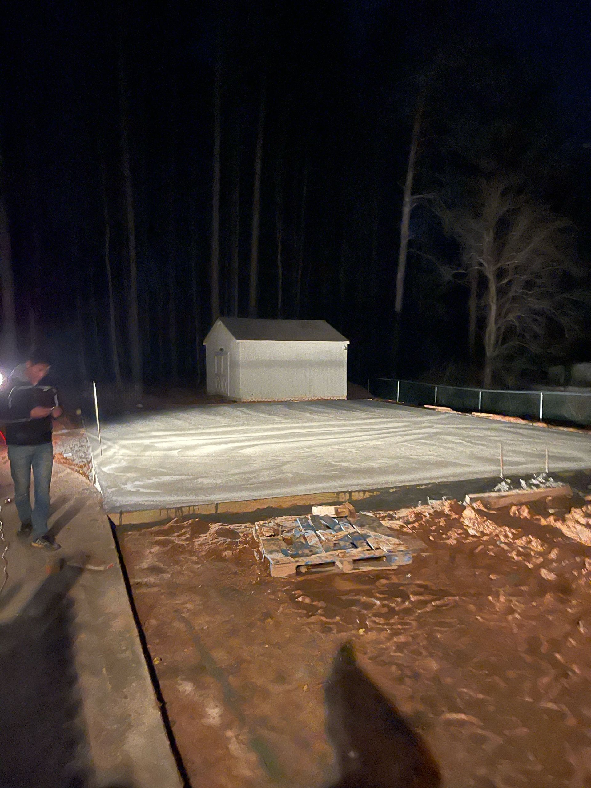 Nighttime construction site with a concrete slab, small shed, and a person standing on the left.