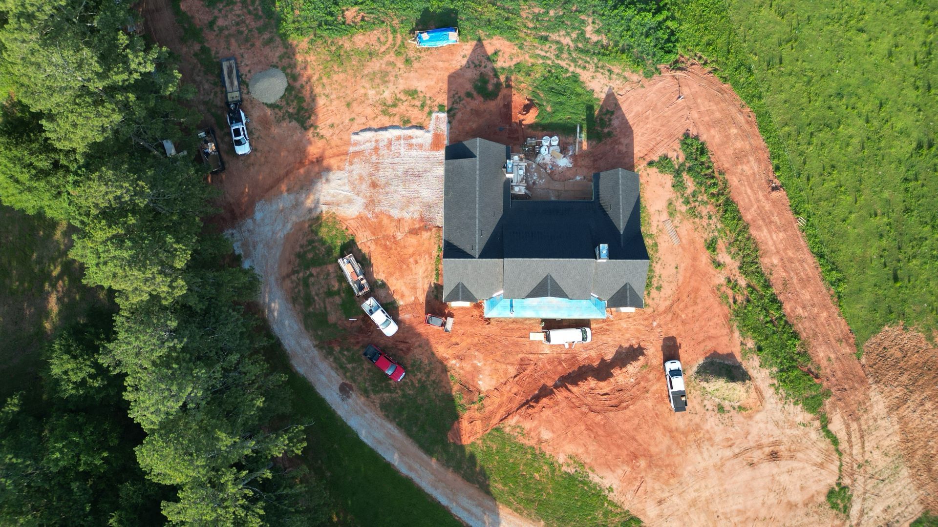 Aerial view of a house under construction with red dirt, trees, and parked vehicles nearby.