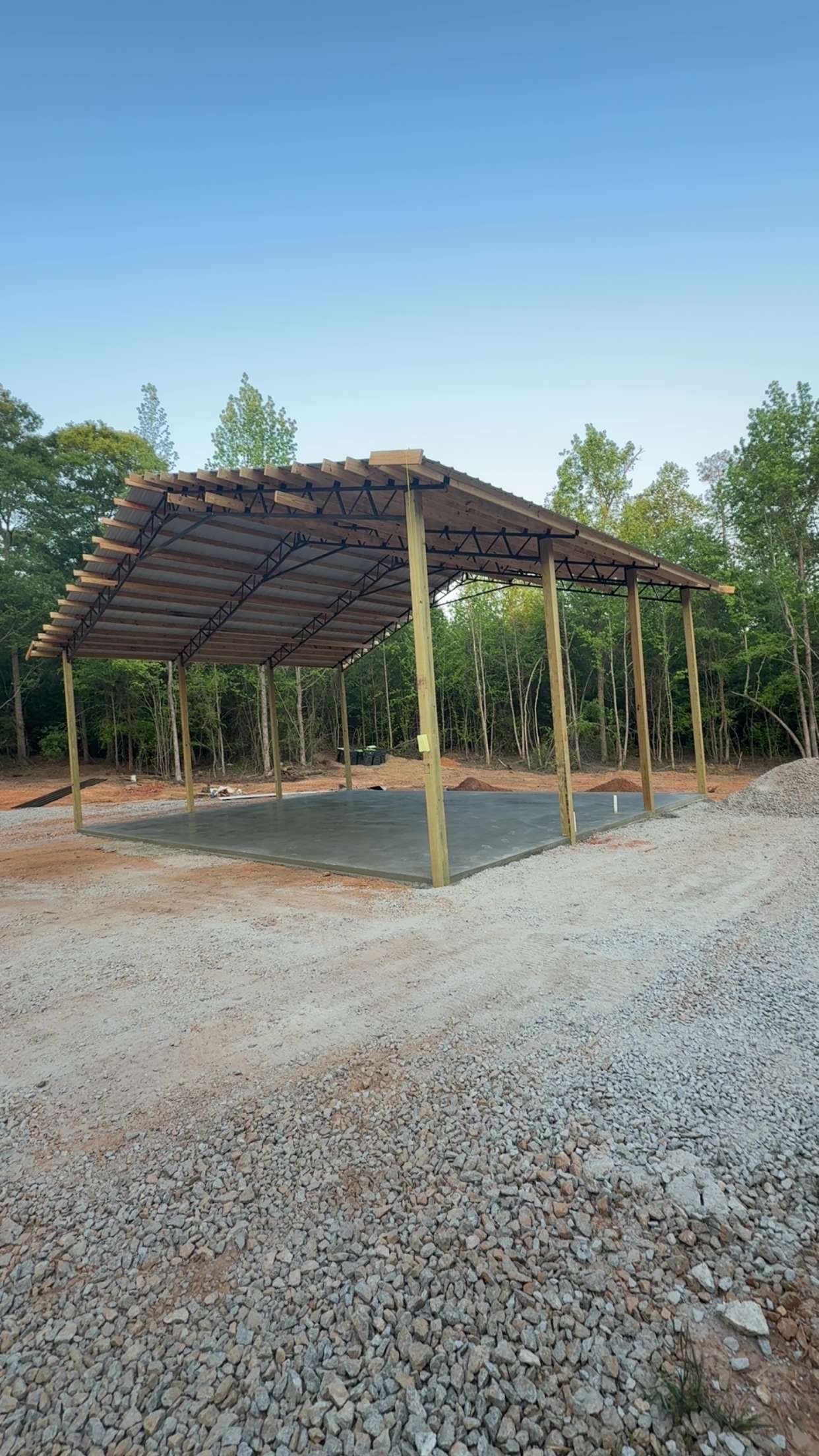 Open-sided bamboo shelter with a thatched roof on a gravel clearing, surrounded by trees and blue sky