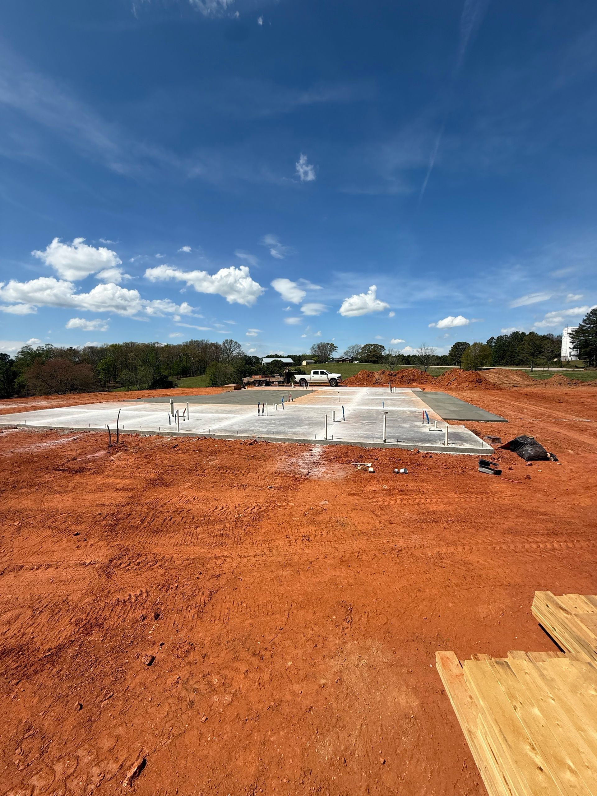 Concrete foundation on red dirt under blue sky with clouds, trees in the distance.