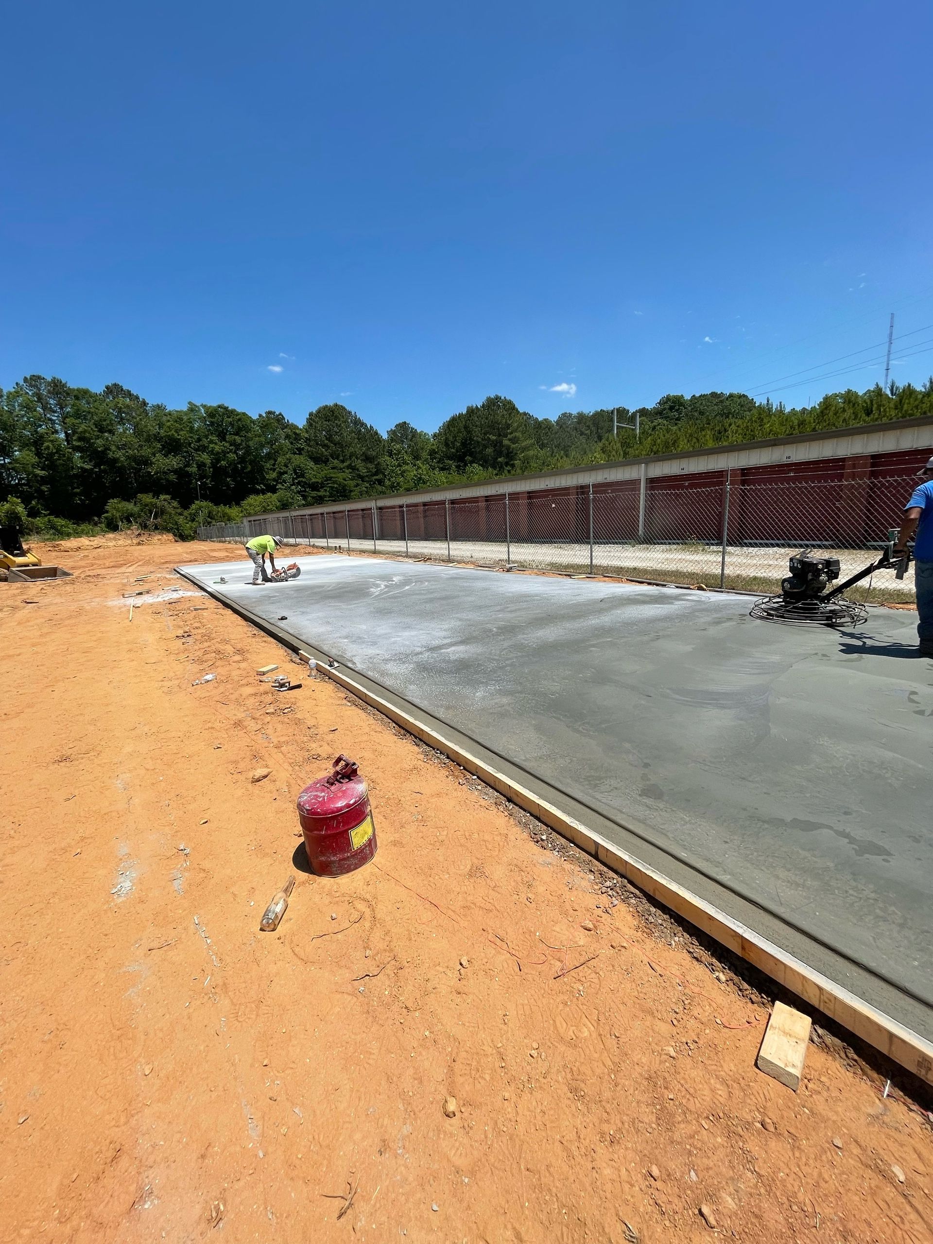 Road construction site with fresh concrete, workers, and an orange dirt roadside under a clear blue sky