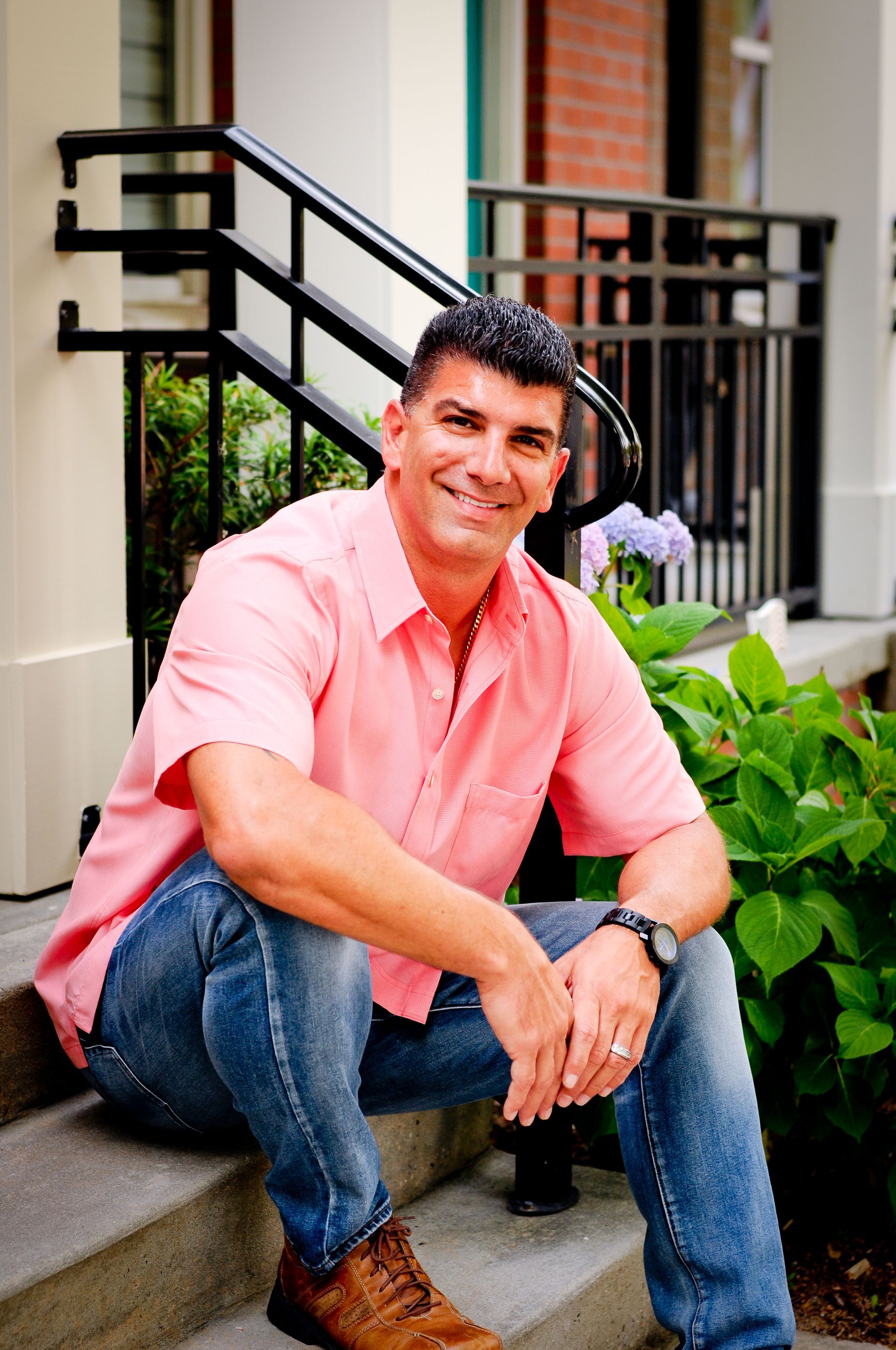 Man sits on steps, wearing pink shirt and jeans, smiling, outdoors.
