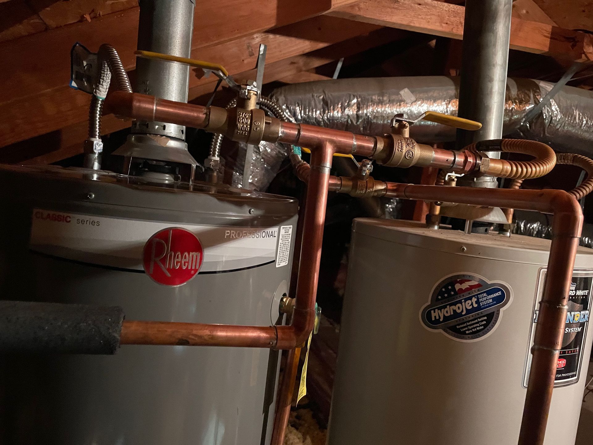 Two water heaters with copper pipes and valves in an attic. One has a red logo, the other a blue logo.