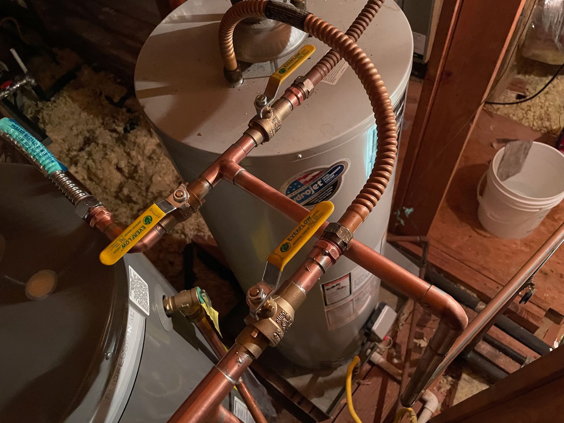 Two water heaters with copper piping and yellow valve handles in an attic setting.