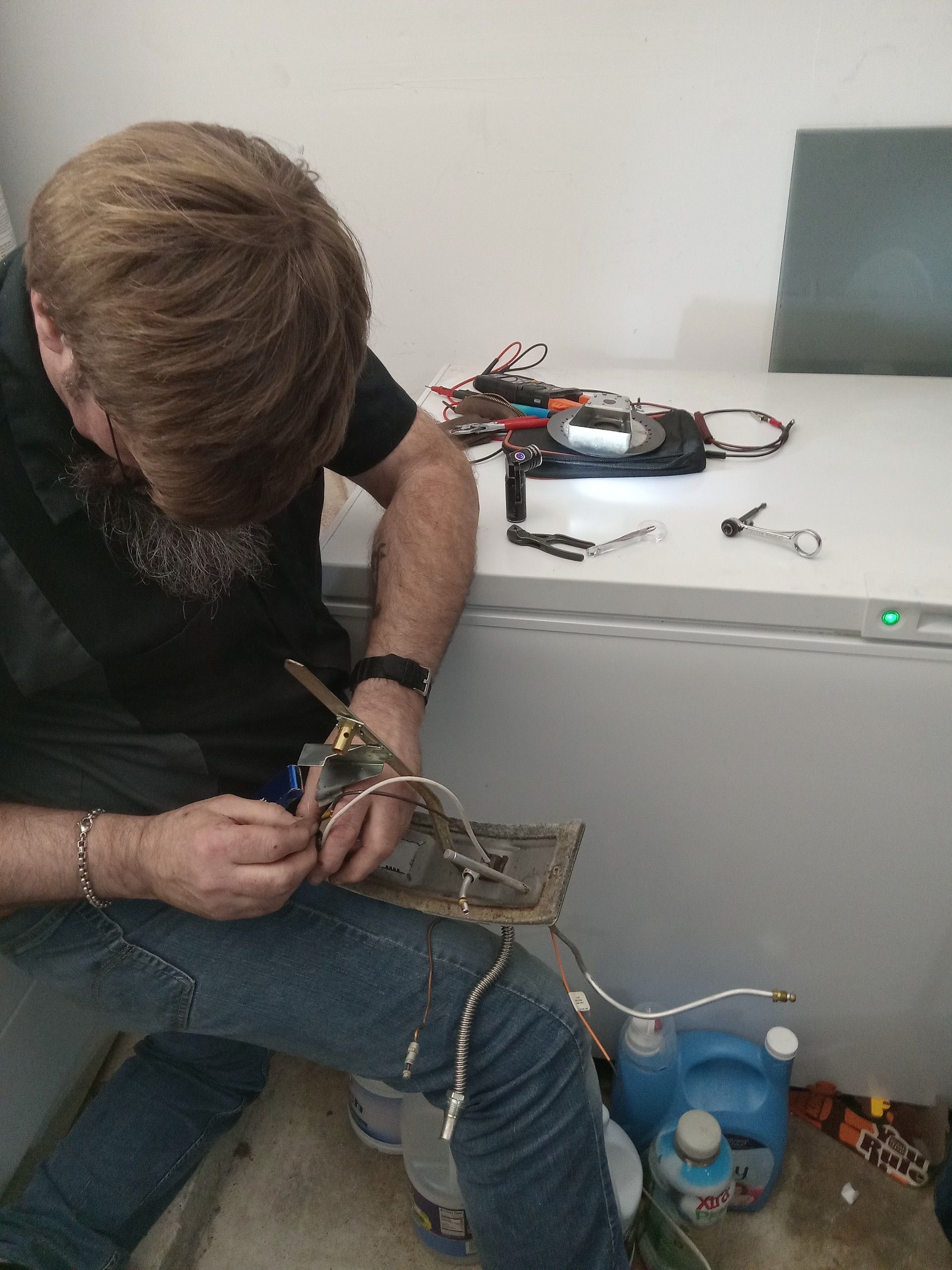 A person working on electronics. They sit near a white freezer, focusing intently on wires and a circuit board.