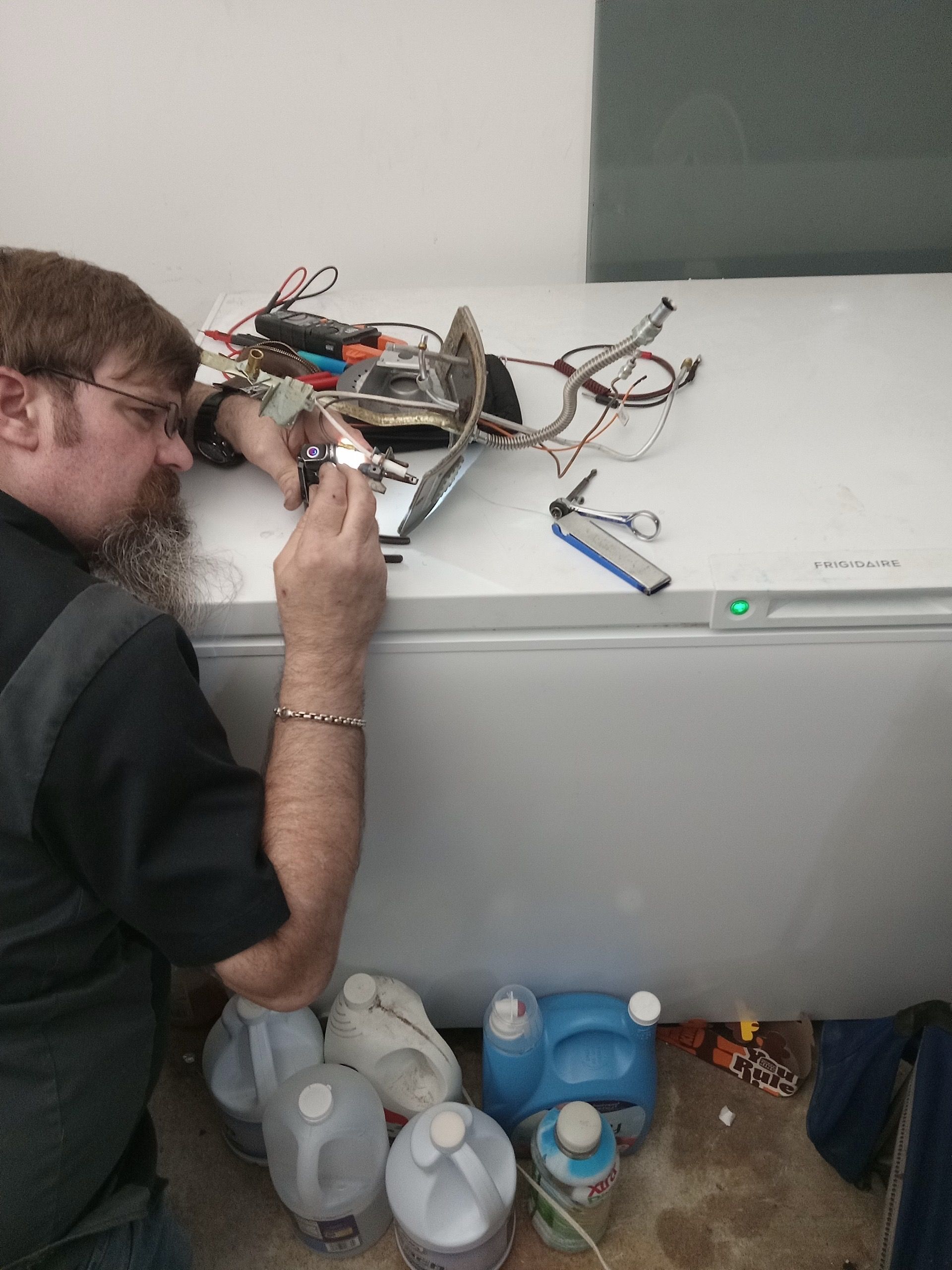 Man with beard working on electrical components atop a white freezer; he’s kneeling on a wet floor with several jugs and bottles around him.