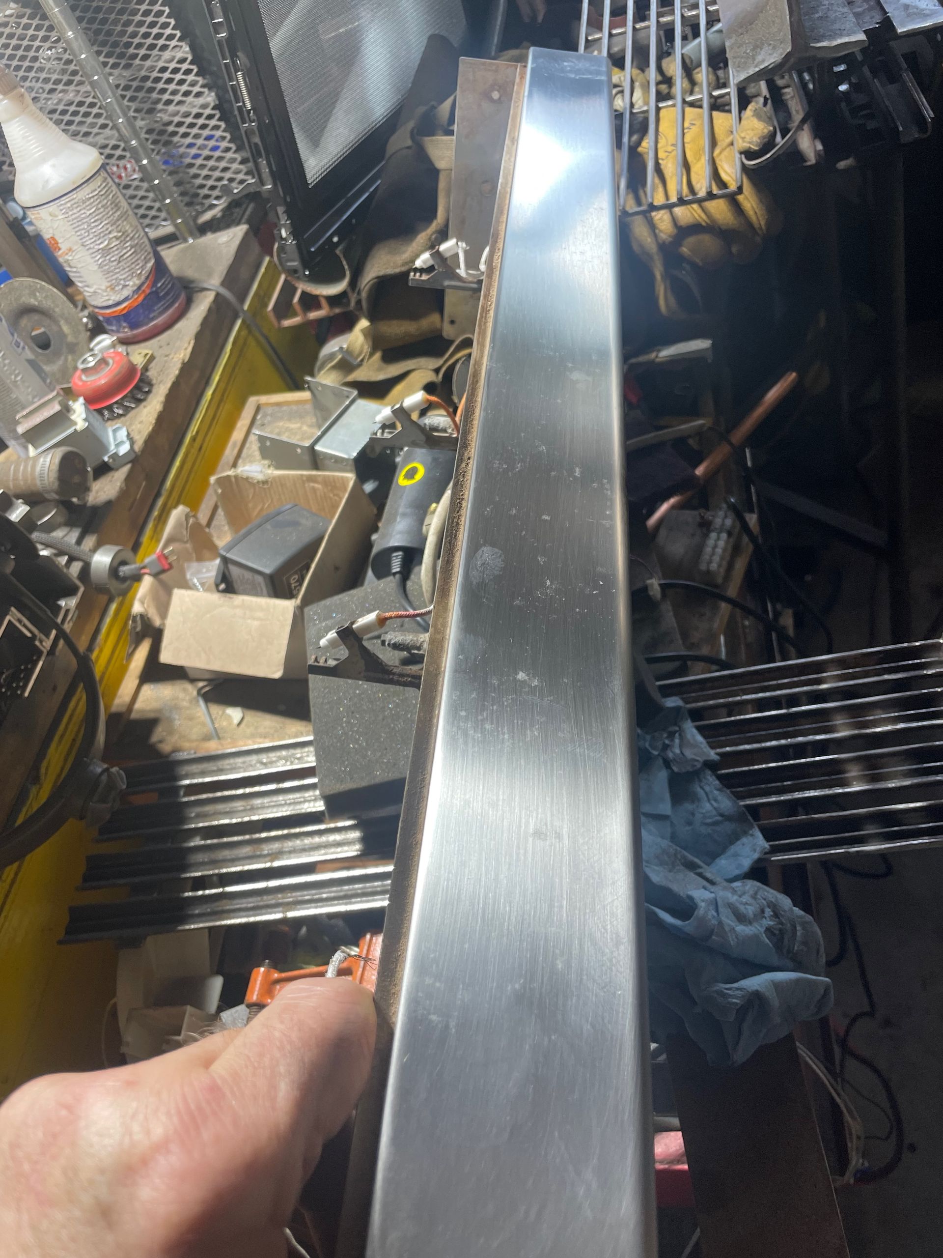 A hand holds a long, shiny, metal piece, possibly part of a larger construction project, in a cluttered workshop.