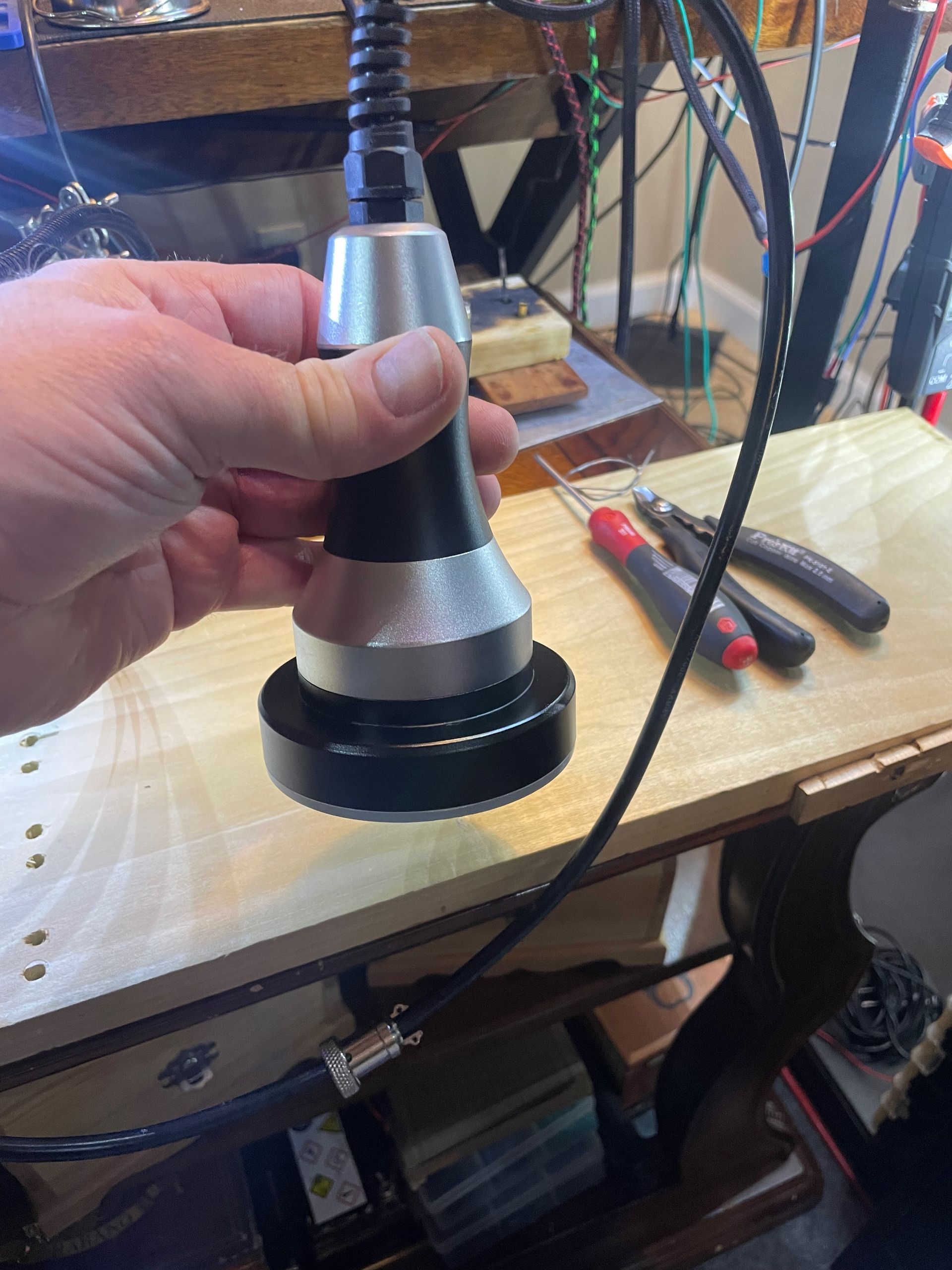 Hand holding a black and silver device with a circular base and a thick cable.  It rests on a wooden surface with tools.