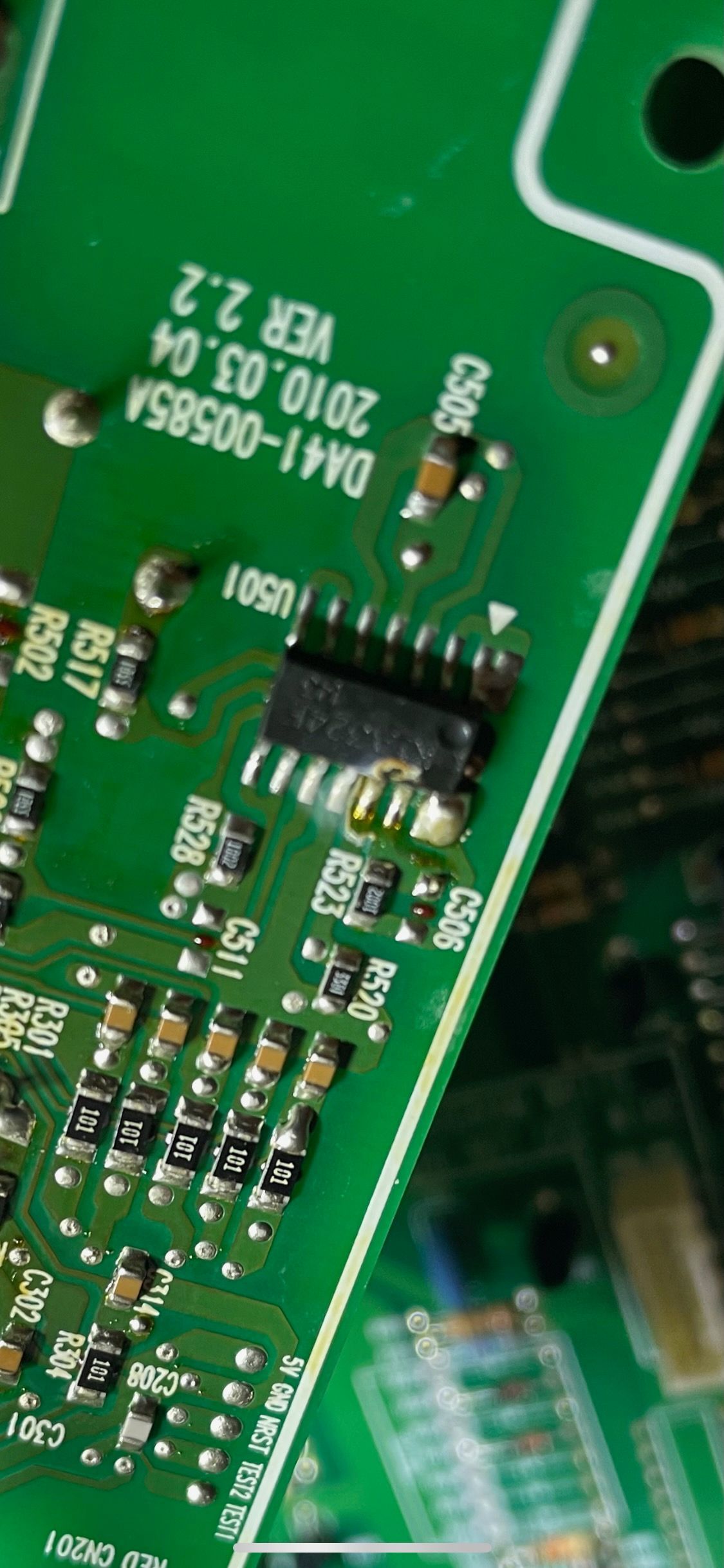 Green circuit board with several electronic components, including a black integrated circuit chip and small brown capacitors.