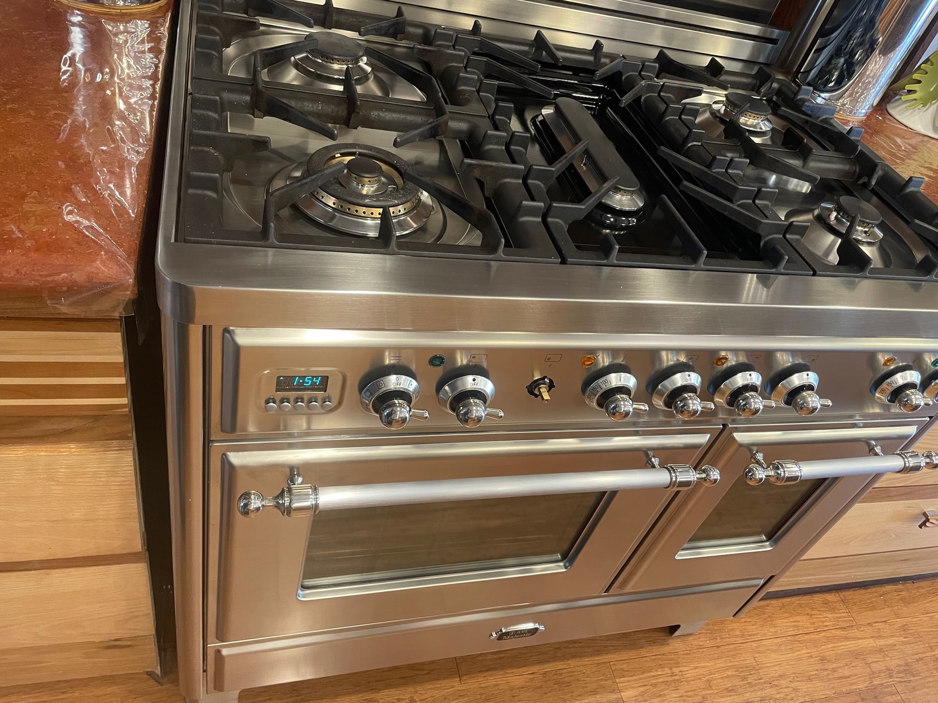 Stainless steel gas range with two ovens and multiple burners, in a kitchen setting. The oven has a digital display and multiple knobs.