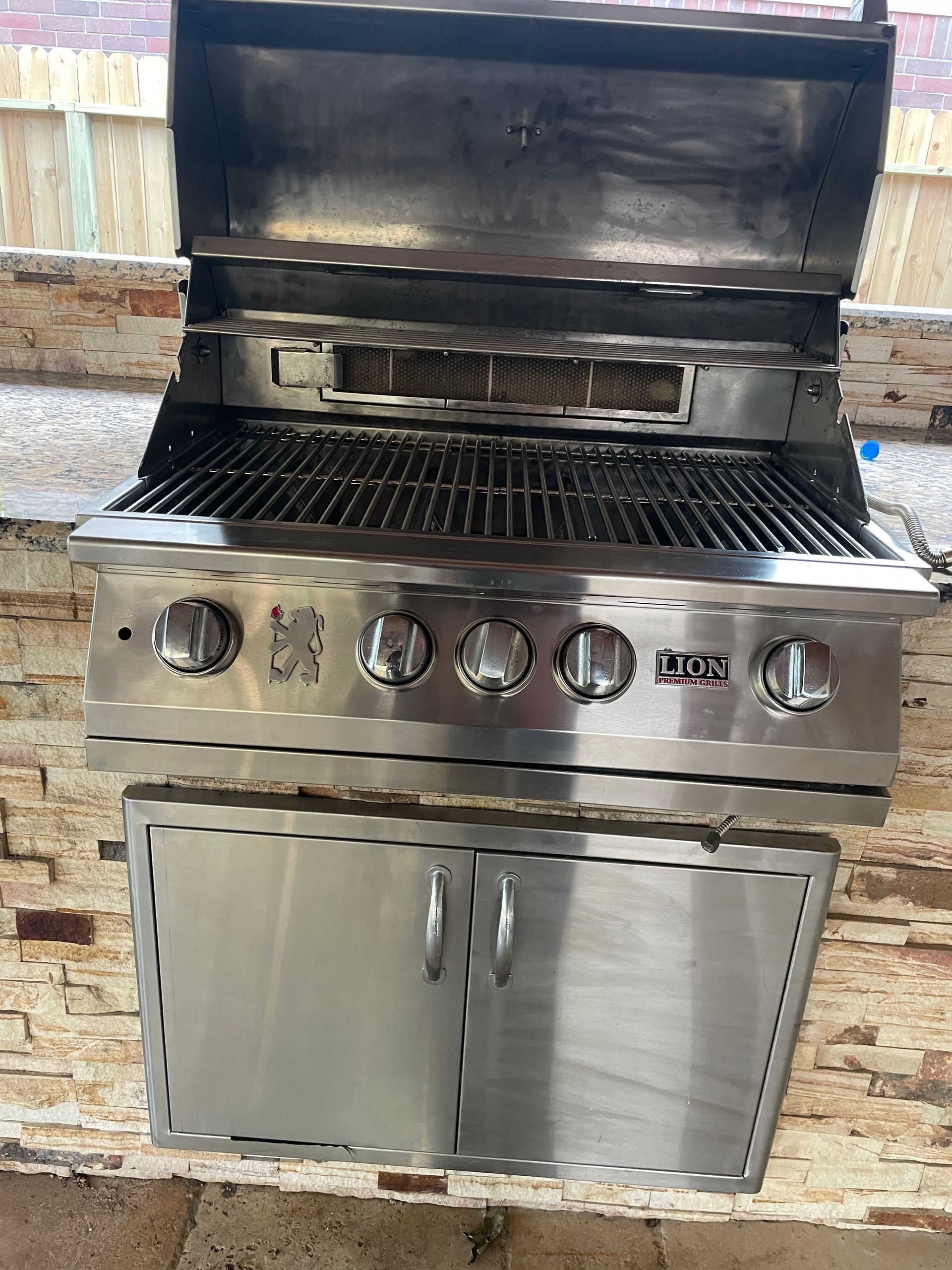 Stainless steel outdoor grill with the lid open, set into a stone facade with cabinet doors below.