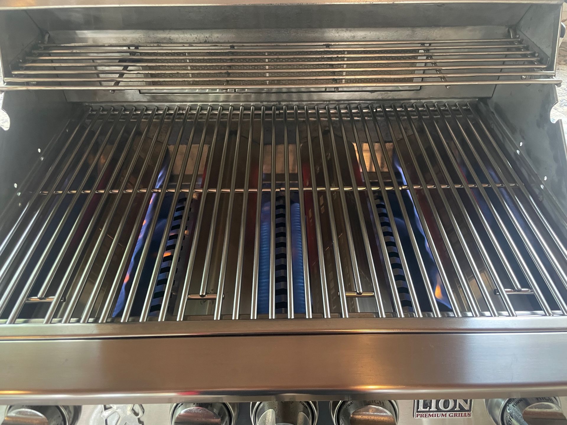 A stainless steel gas grill with blue flames rising from the burners. The grill grates are visible.