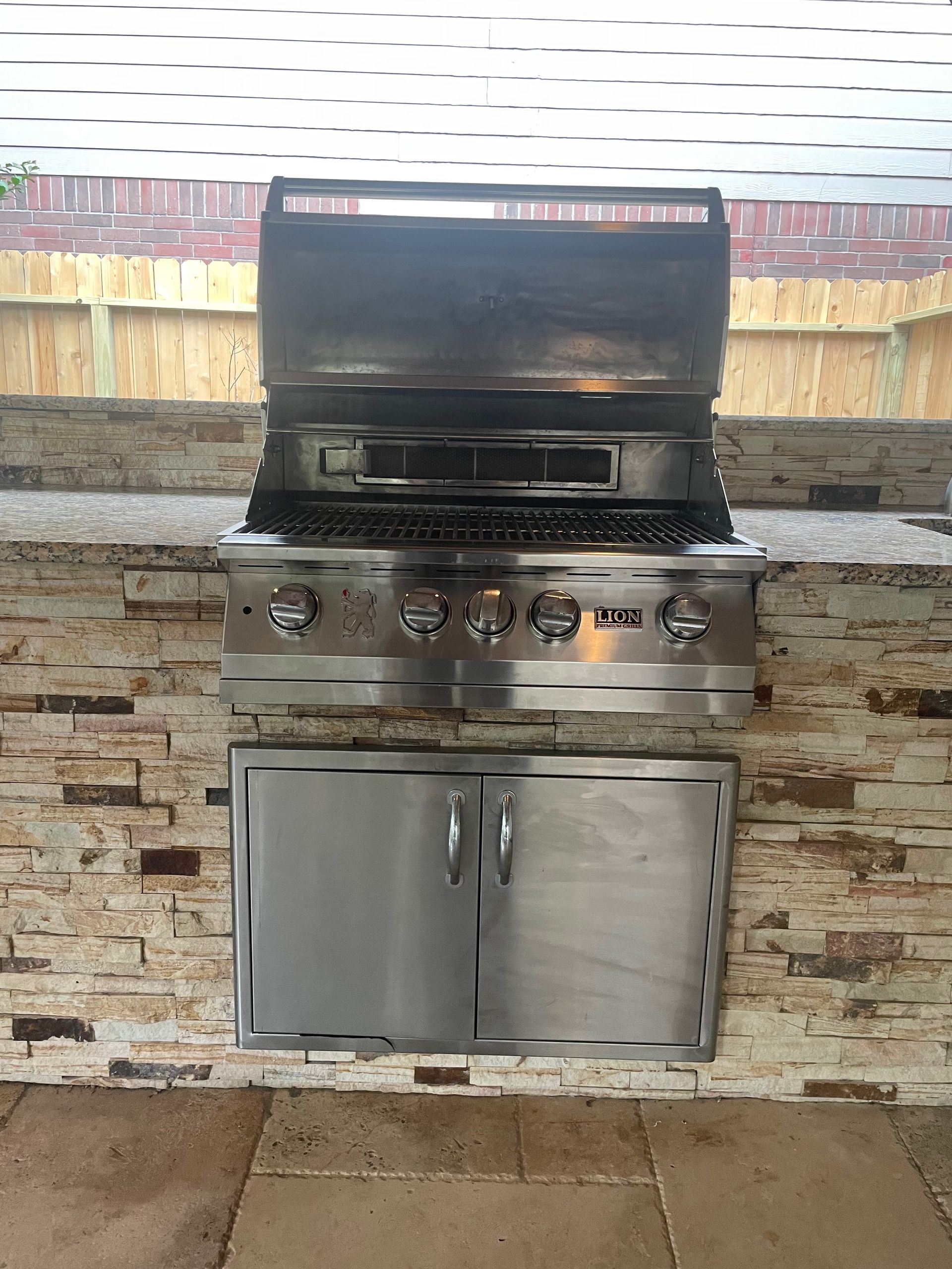 A stainless steel outdoor grill built into a stone facade. Below the grill is a stainless steel cabinet.