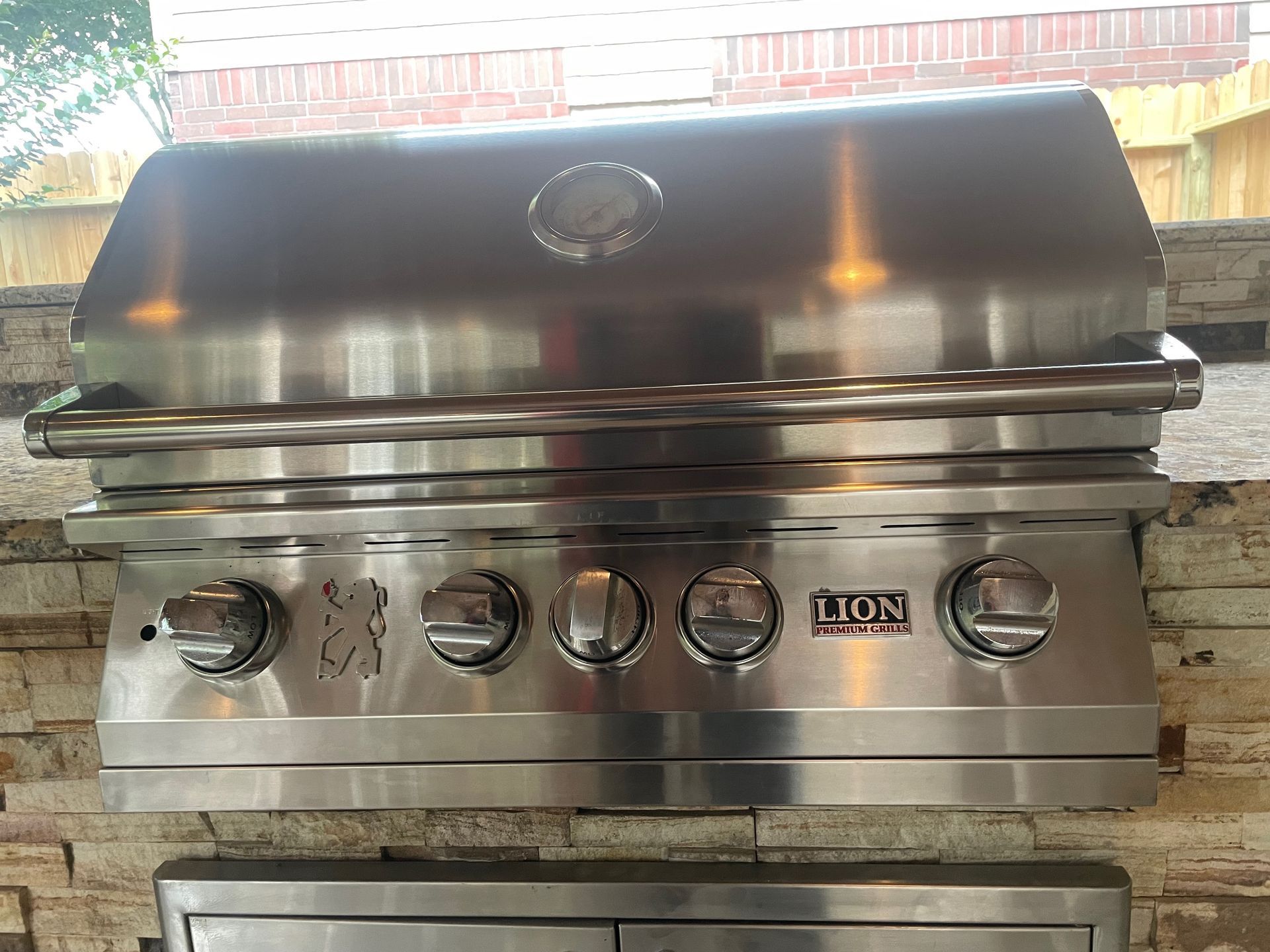 Stainless steel Lion gas grill built into a brick outdoor kitchen counter. The grill has five control knobs and a temperature gauge.