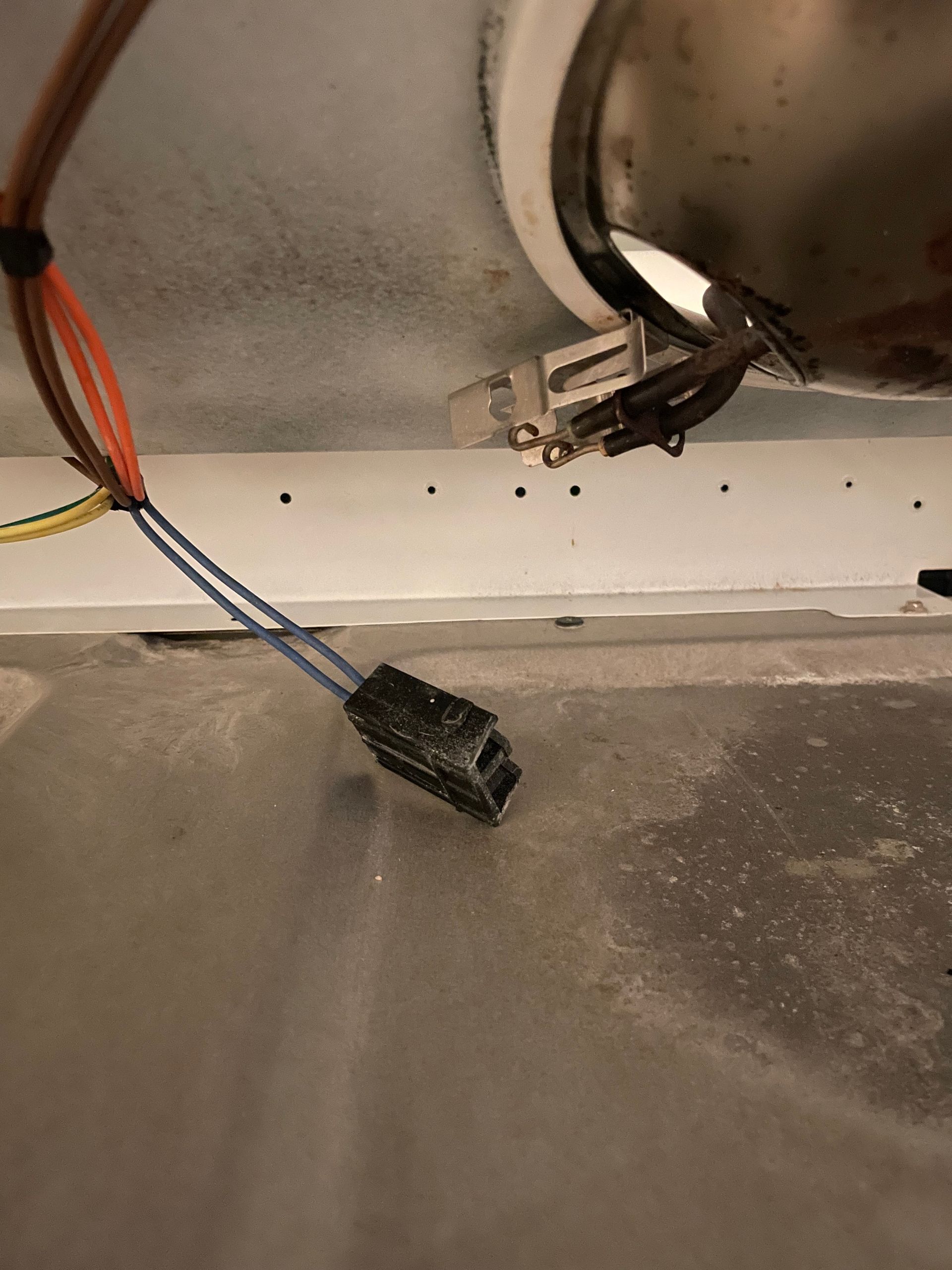 Inside a white dryer, a black electrical connector with blue wires is visible. Other wires and a metal component are in the background.