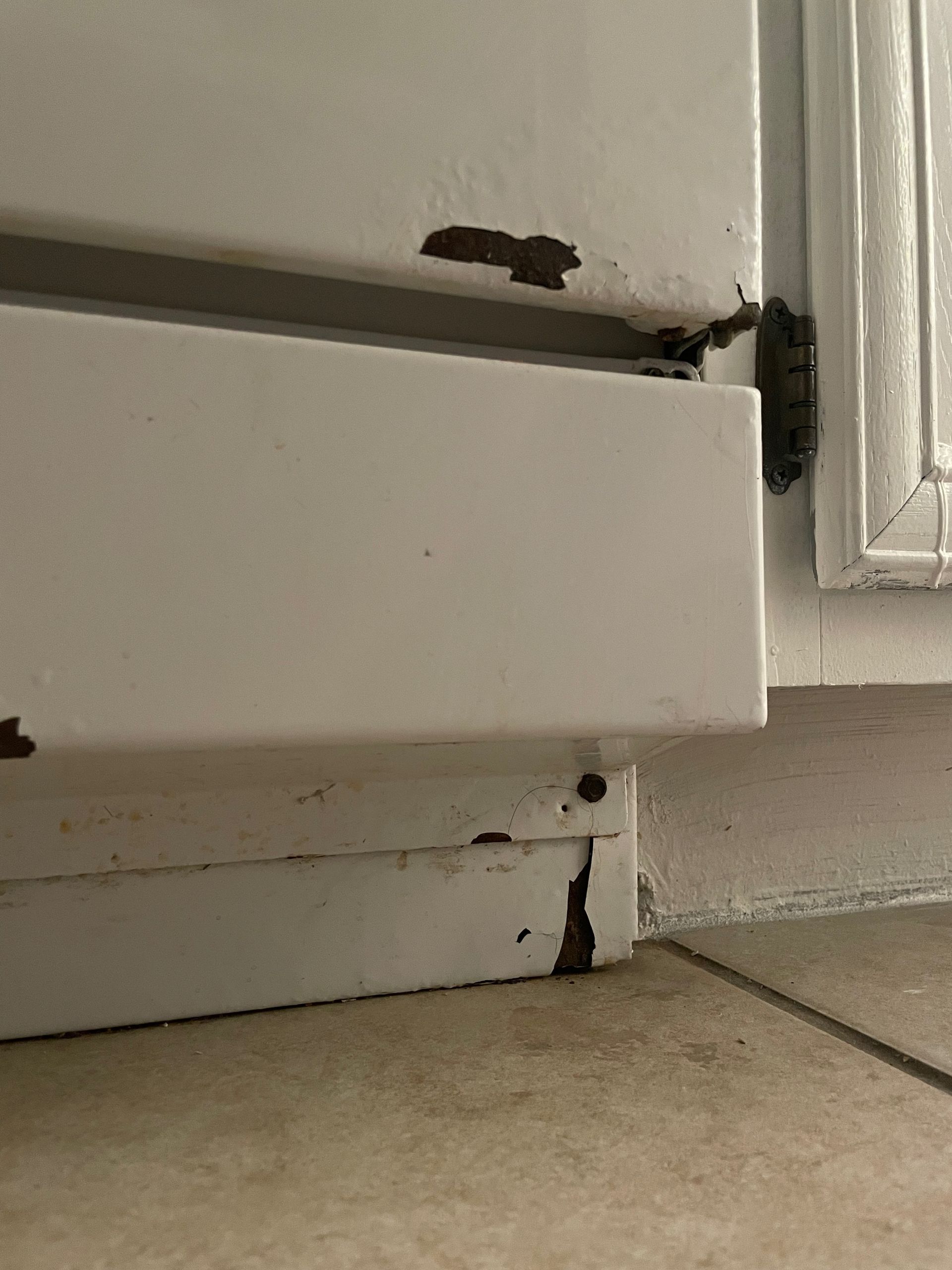 Close-up of a white metal door frame with peeling paint, likely from water damage, near a tiled floor.