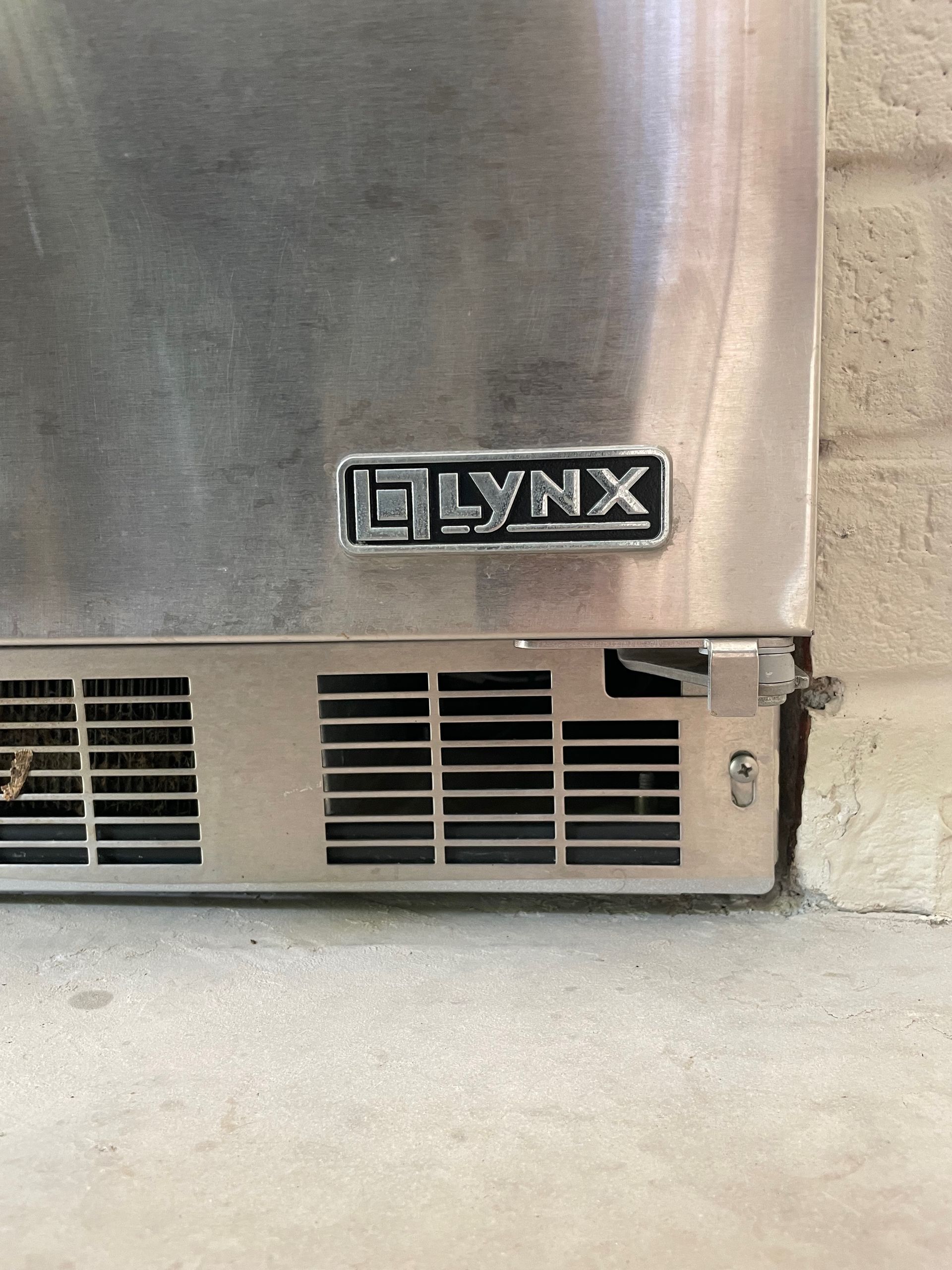 Close-up of a stainless steel Lynx outdoor refrigerator. The logo is visible, along with the vent and a small door hinge.