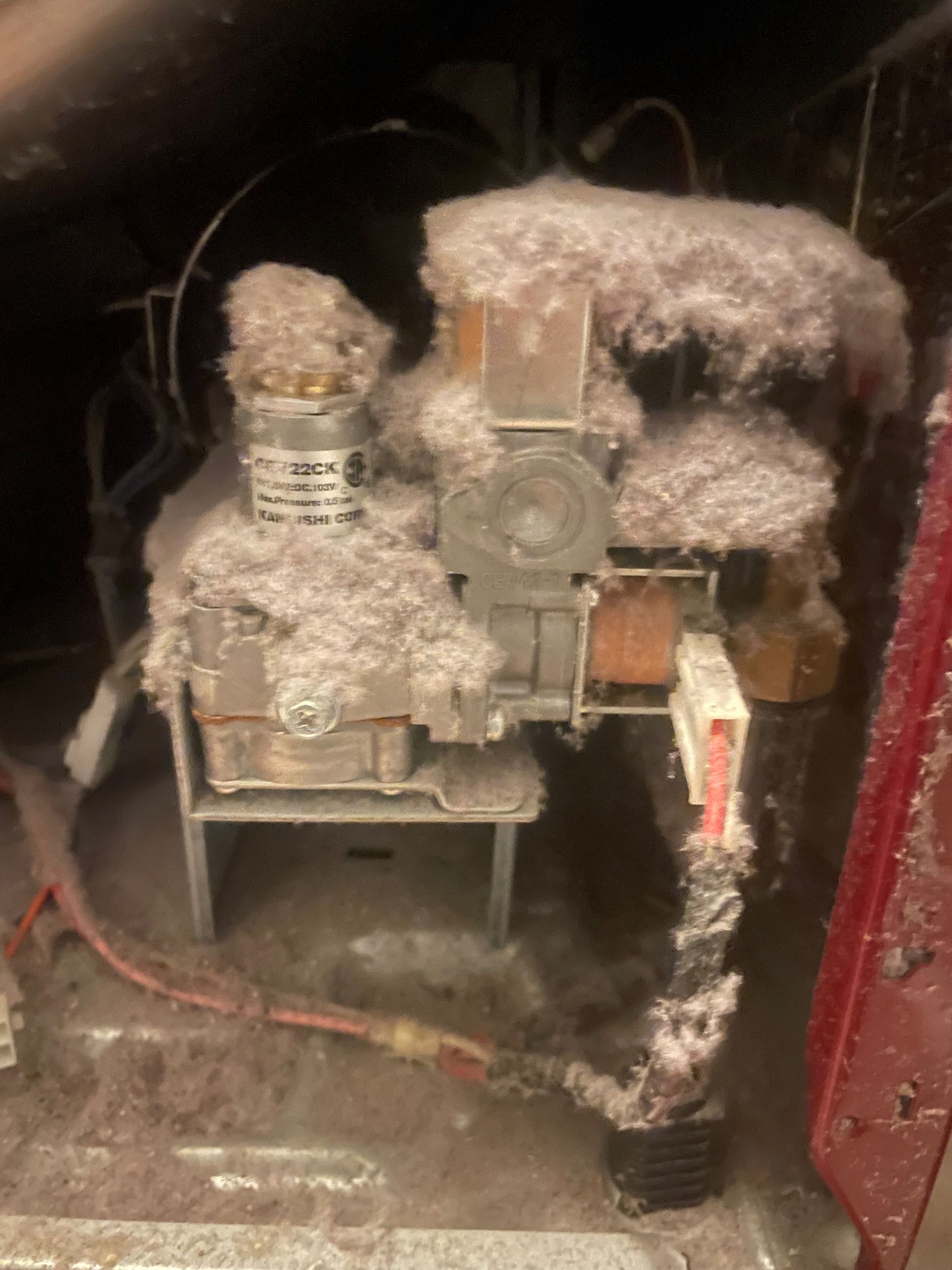 Heavily dust-covered furnace components. Thick accumulation of gray lint coats the metal parts and wiring inside a reddish housing.
